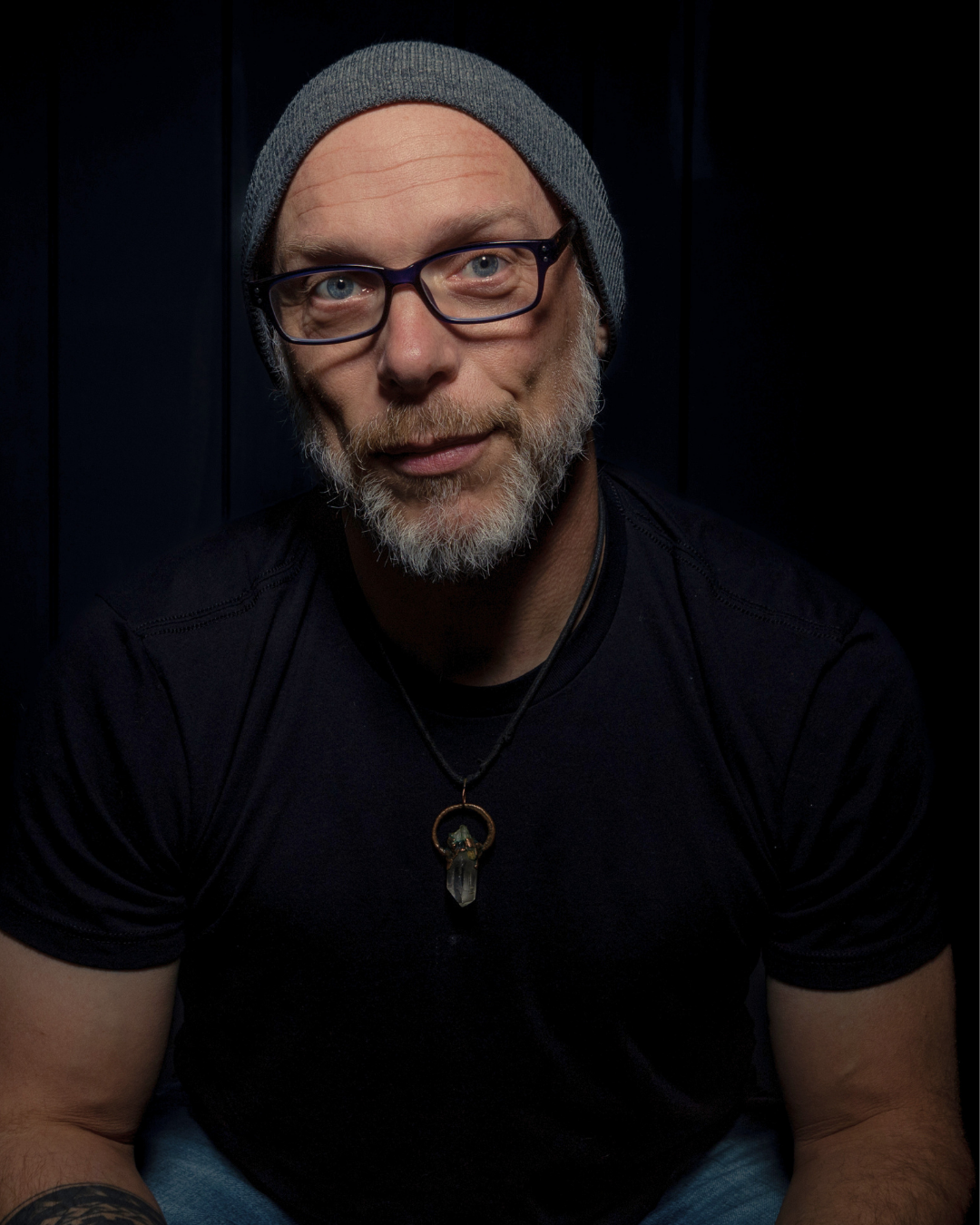 Portrait of a man Mahoe Movement mindset coach, speaker, and meditation teacher Joe Longo with glasses, wearing a gray beanie and a necklace, in a dark setting.