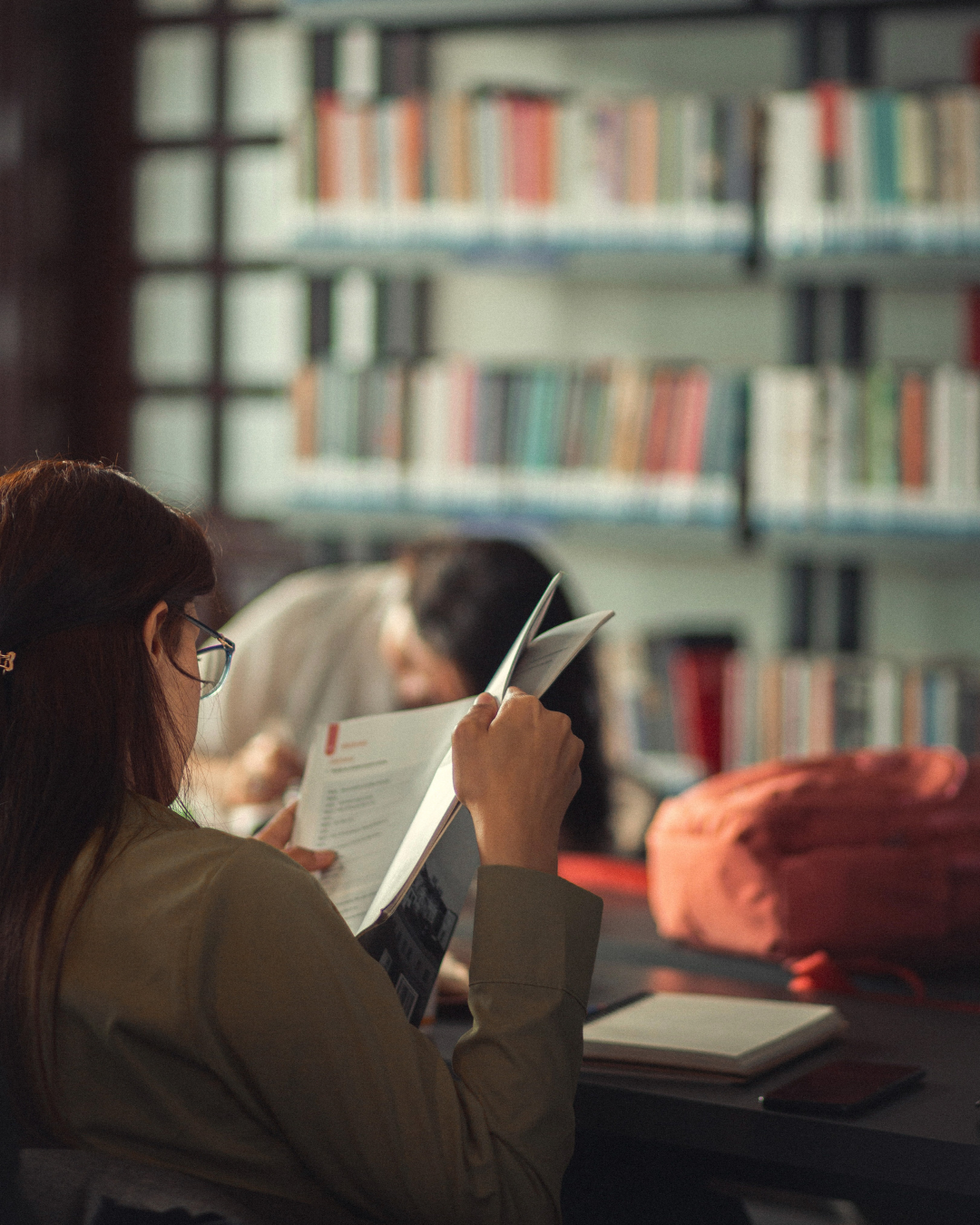 Women studying or reading at a table in a university library with bookshelves in the background.