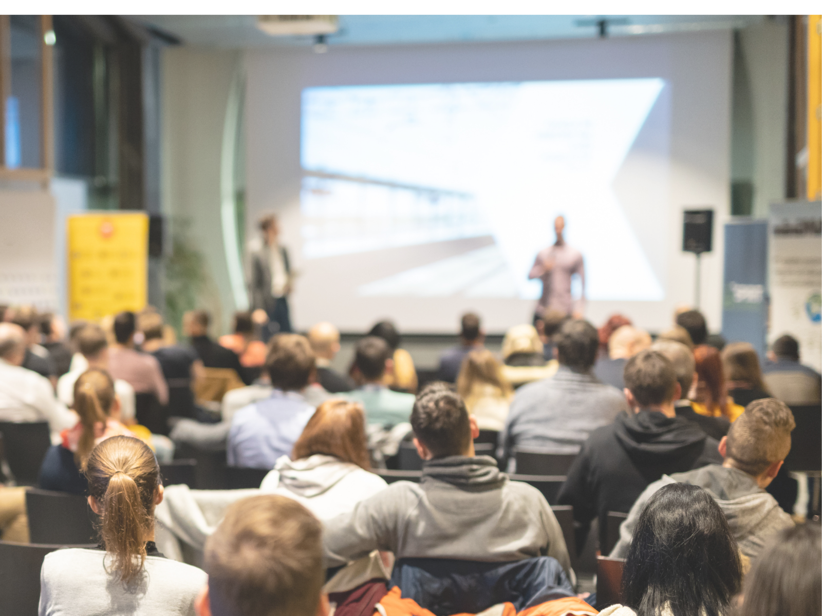 Audience attending a conference with two speakers presenting on stage with a large screen in the background.