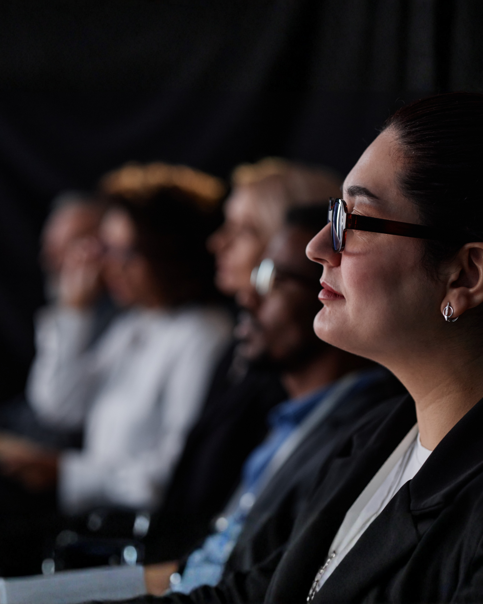 A side profile of a woman with glasses, sitting in a row with others, during what appears to be a formal event or conference.