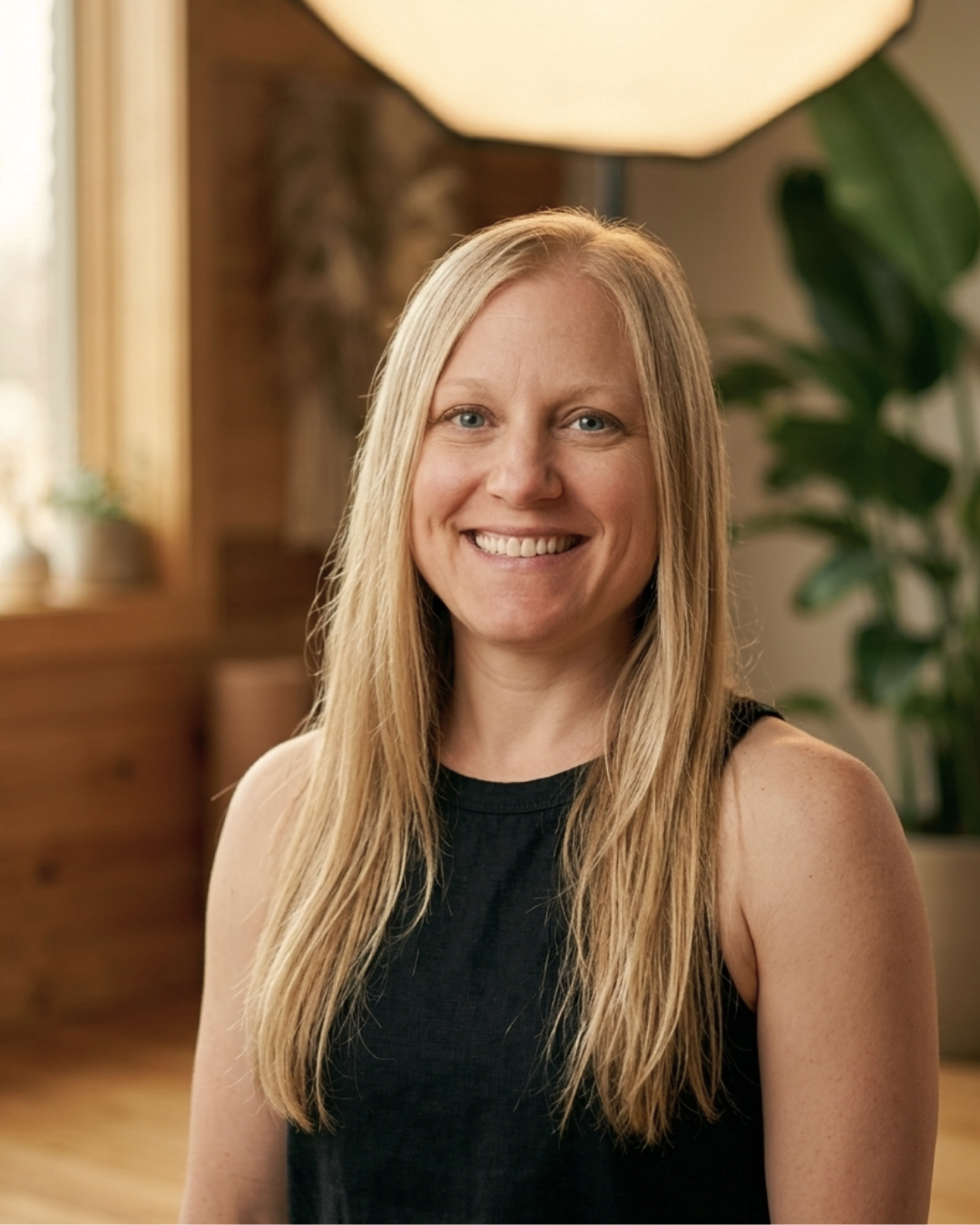 A smiling woman with long blonde hair Mahoe Movement nutrition and movement instructor Kelly Pauly in a black sleeveless top, standing indoors with a wooden wall, large window, and green plant in the background.