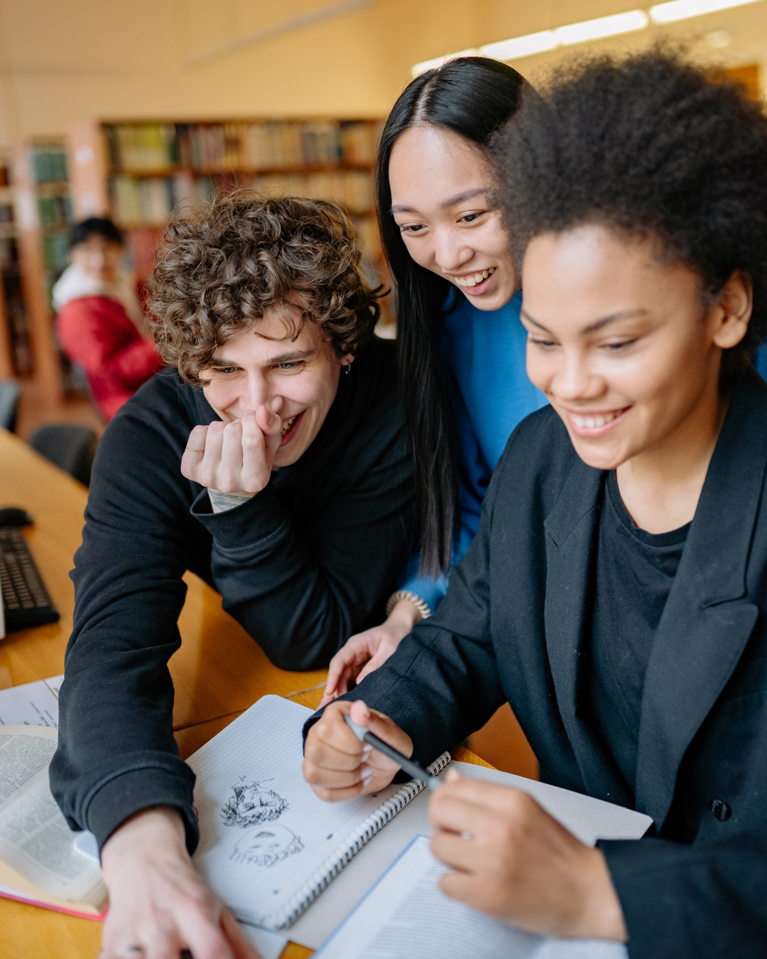 Three students gathered around a desk, smiling and looking at a drawing in a notebook, with bookshelves in the background.