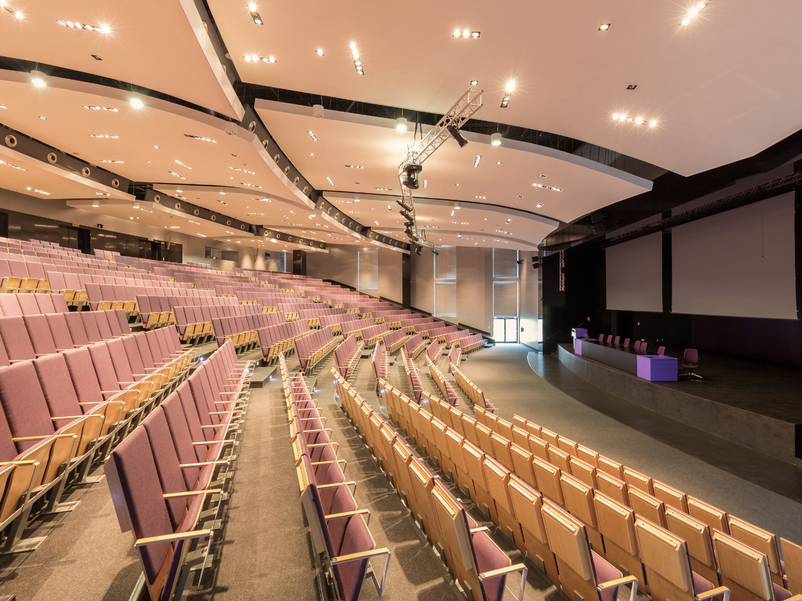 Empty auditorium with rows of purple and beige seats, large screens on stage, and stage lighting.