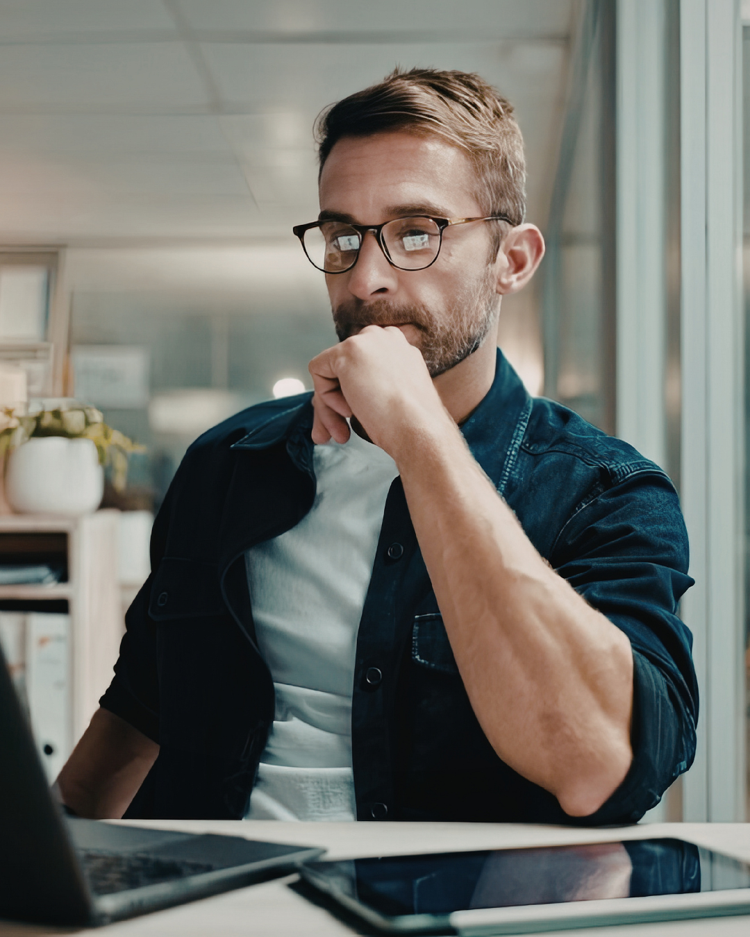 A man with glasses, beard, and short brown hair, wearing a denim jacket over a white shirt, sitting at a desk in an office, looking at a computer screen with a thoughtful expression.