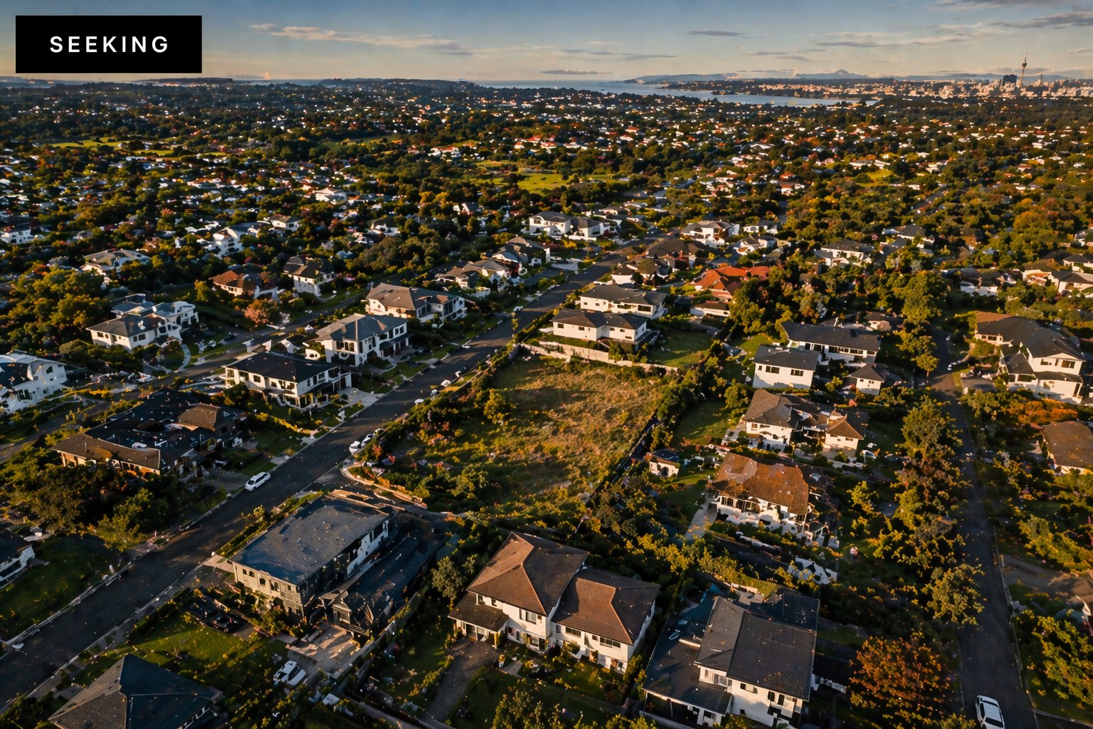 Aerial view of a suburban neighborhood with numerous houses, trees, and streets, taken during sunset with city skyline and water in the distance.