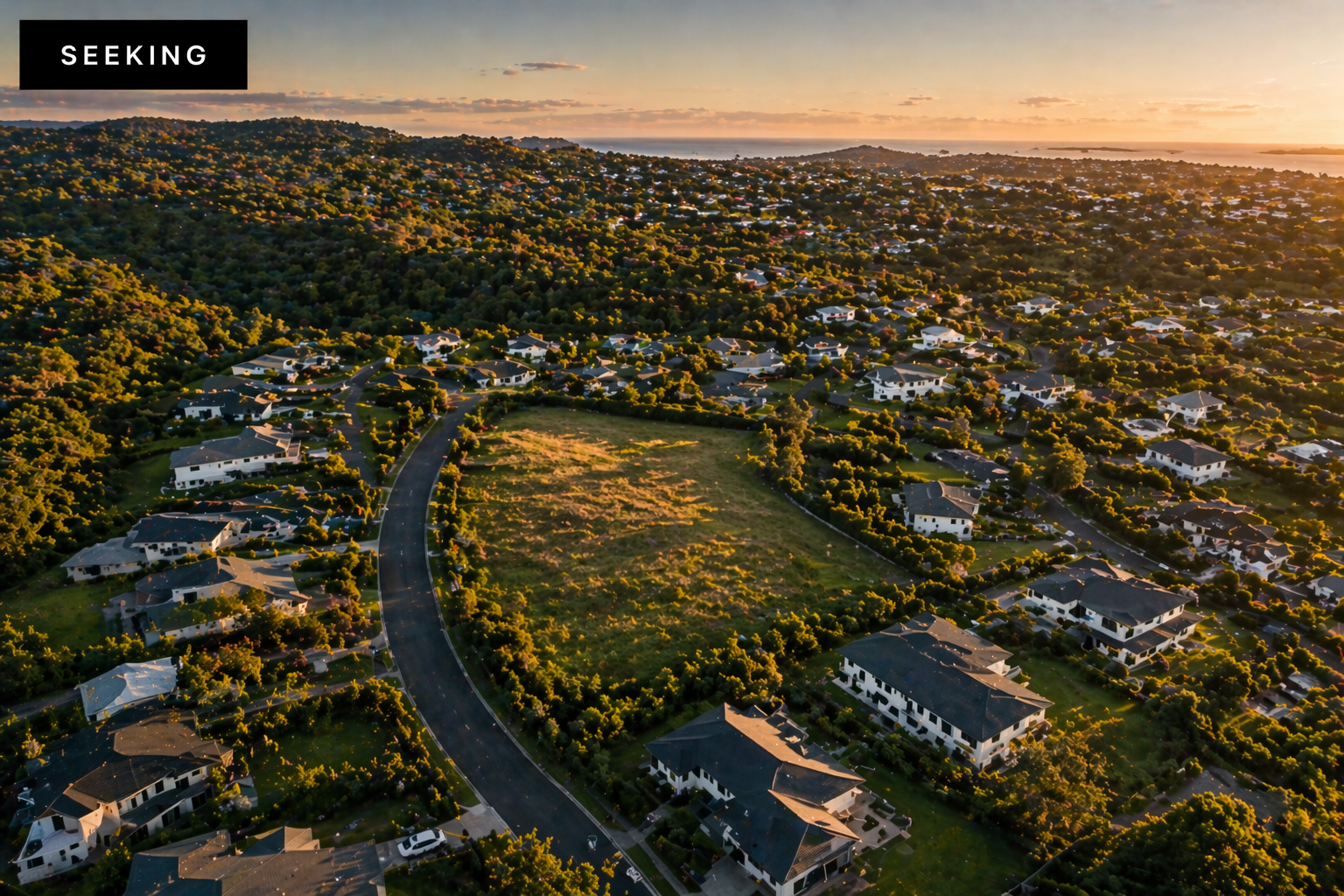 Aerial view of a suburban neighborhood with houses, tree-lined streets, and a large open grassy area during sunset.