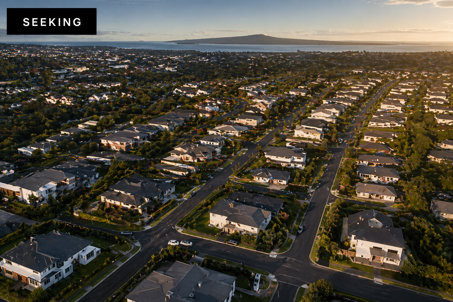Aerial view of a suburban neighborhood with many houses, trees, and roads during sunset, overlooking a body of water and distant hills.
