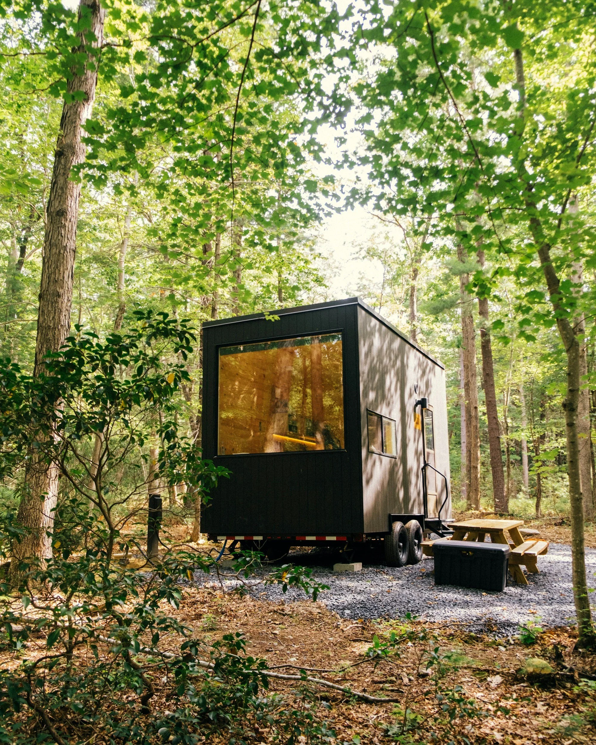 A black tiny house on wheels parked in a wooded area with green trees, a small wooden picnic table, and a storage container nearby.