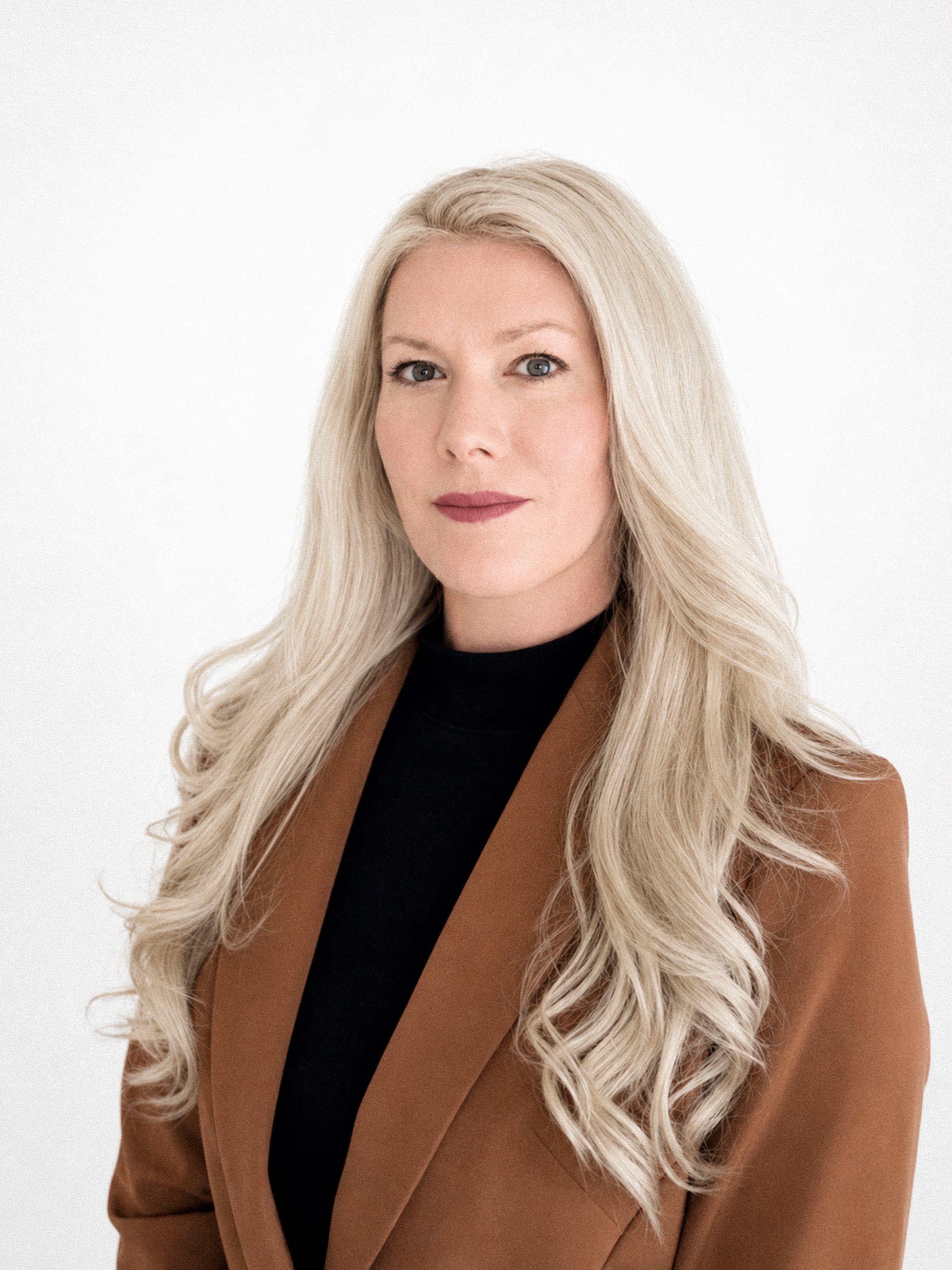 A woman with long blonde hair, blue eyes, and fair skin, wearing a black top and a brown blazer, posing against a plain white background.