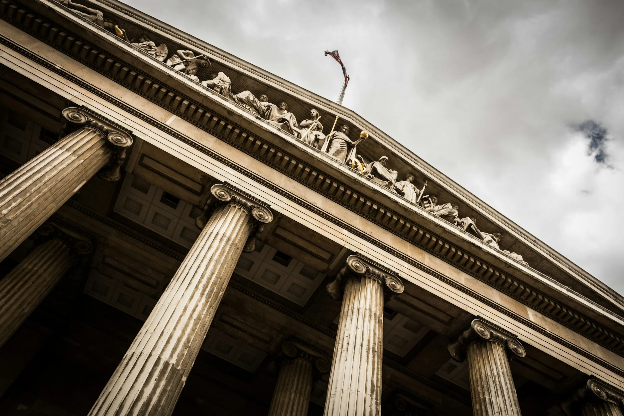 Low-angle view of a classical building with large columns and detailed sculptures on the upper frieze, under a cloudy sky.