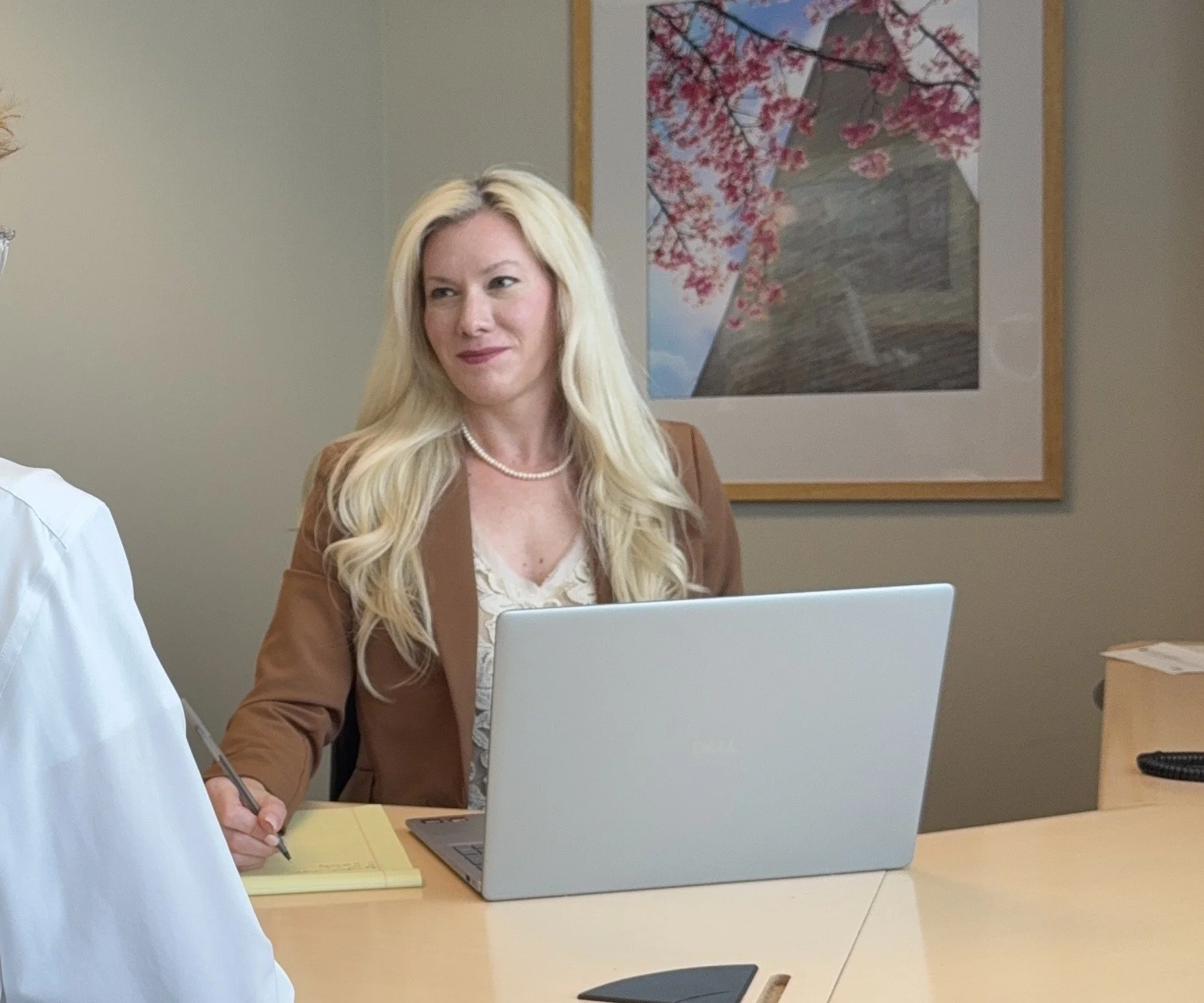 A woman with long blonde hair, wearing a pearl necklace and brown blazer, sitting at a desk with a laptop, talking to a person wearing a white shirt who is holding a pen over yellow notepad. There is a framed picture of cherry blossoms on the wall behind her.