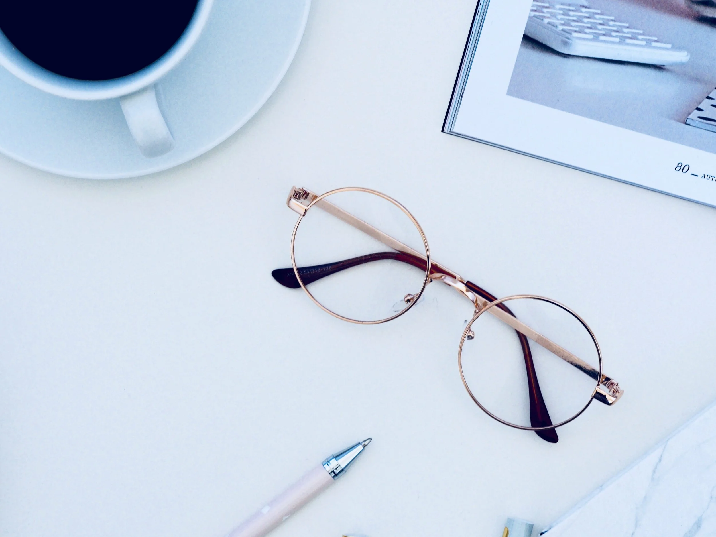 A white desk with a white coffee cup on a saucer, a pair of rose gold eyeglasses, a white pen, and part of a notebook with a visible page. In the background, there is a computer keyboard and a framed picture.