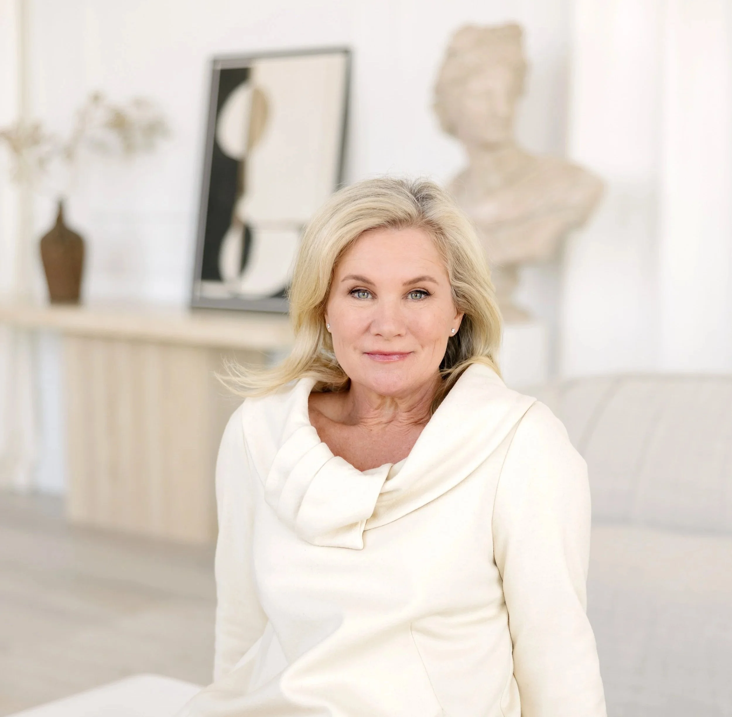 Close-up of a smiling woman with blonde hair and light skin, wearing a white top, against a blurred neutral indoor background with art pieces and a bust sculpture.