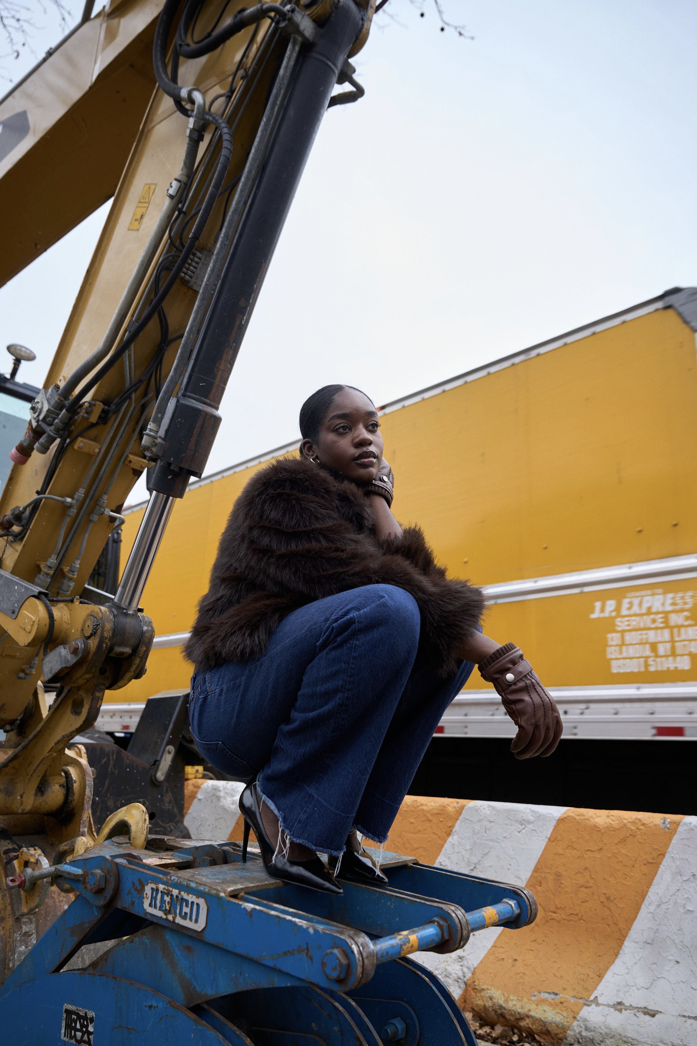 A young woman with dark hair and a serious expression, dressed in a fur jacket, blue jeans, and leather gloves, sitting on construction equipment in front of a yellow truck during daytime.