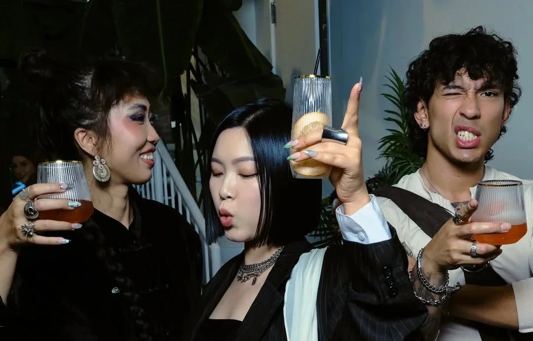 Three young adults holding drinks at a social gathering, making playful and expressive faces.