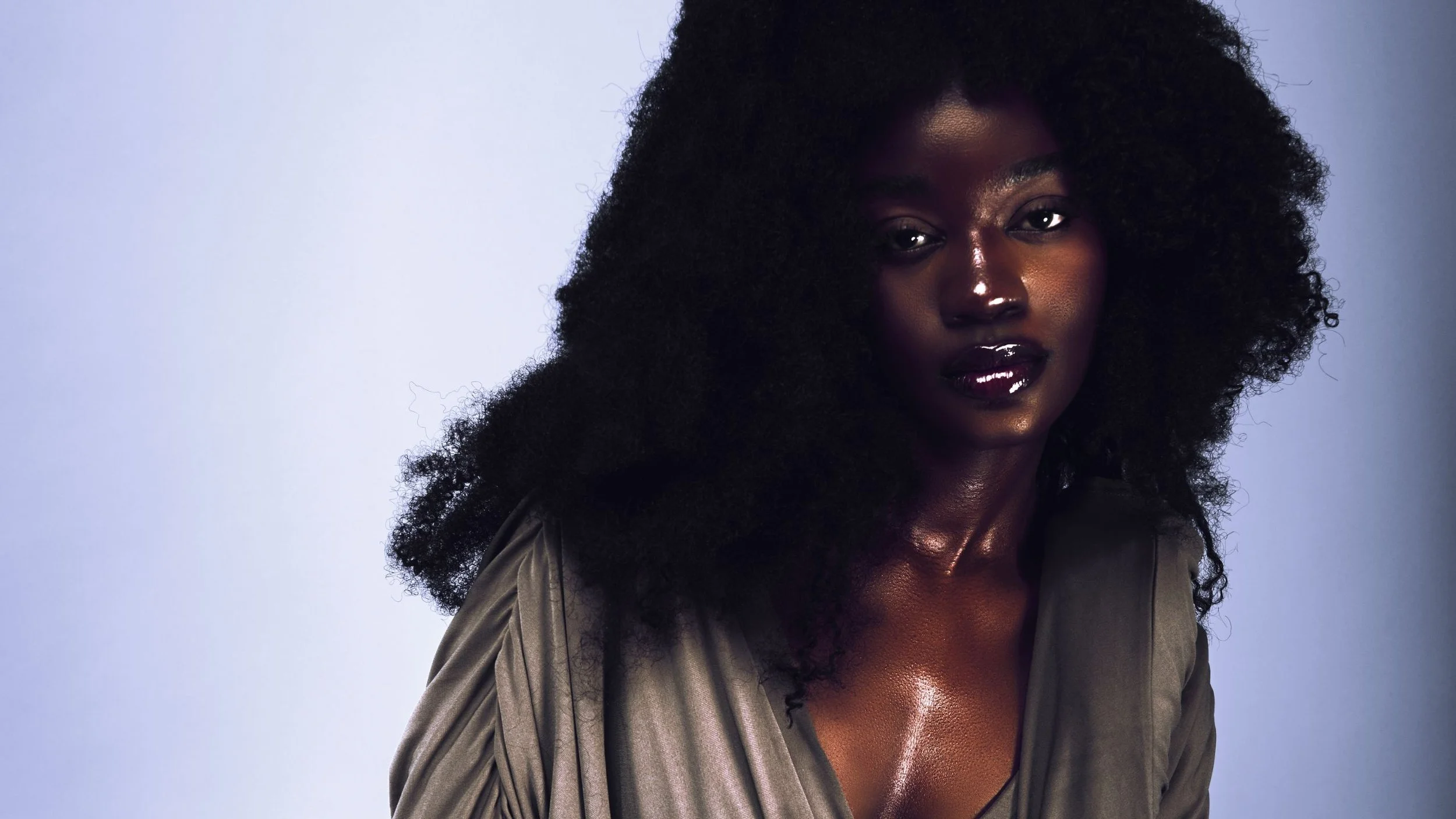 A portrait of a woman with dark skin, voluminous curly black hair, wearing a dark jacket, looking at the camera with a neutral expression against a light blue background.