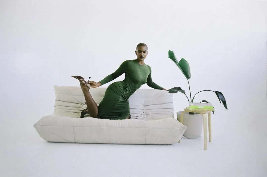 A woman in a green dress is lying face down on a white sofa, holding a high heel shoe in one hand and leaning on a small table with a plant and some books. The background is plain white.
