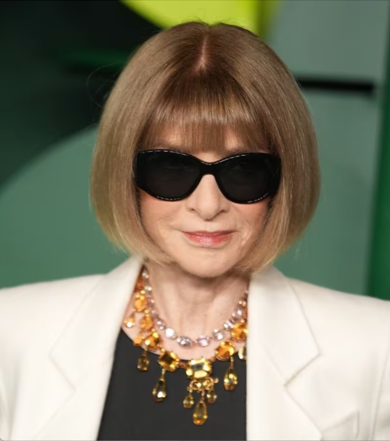 A woman with a bob haircut wearing dark sunglasses, a white blazer, a black top, and a gold and gemstone necklace.