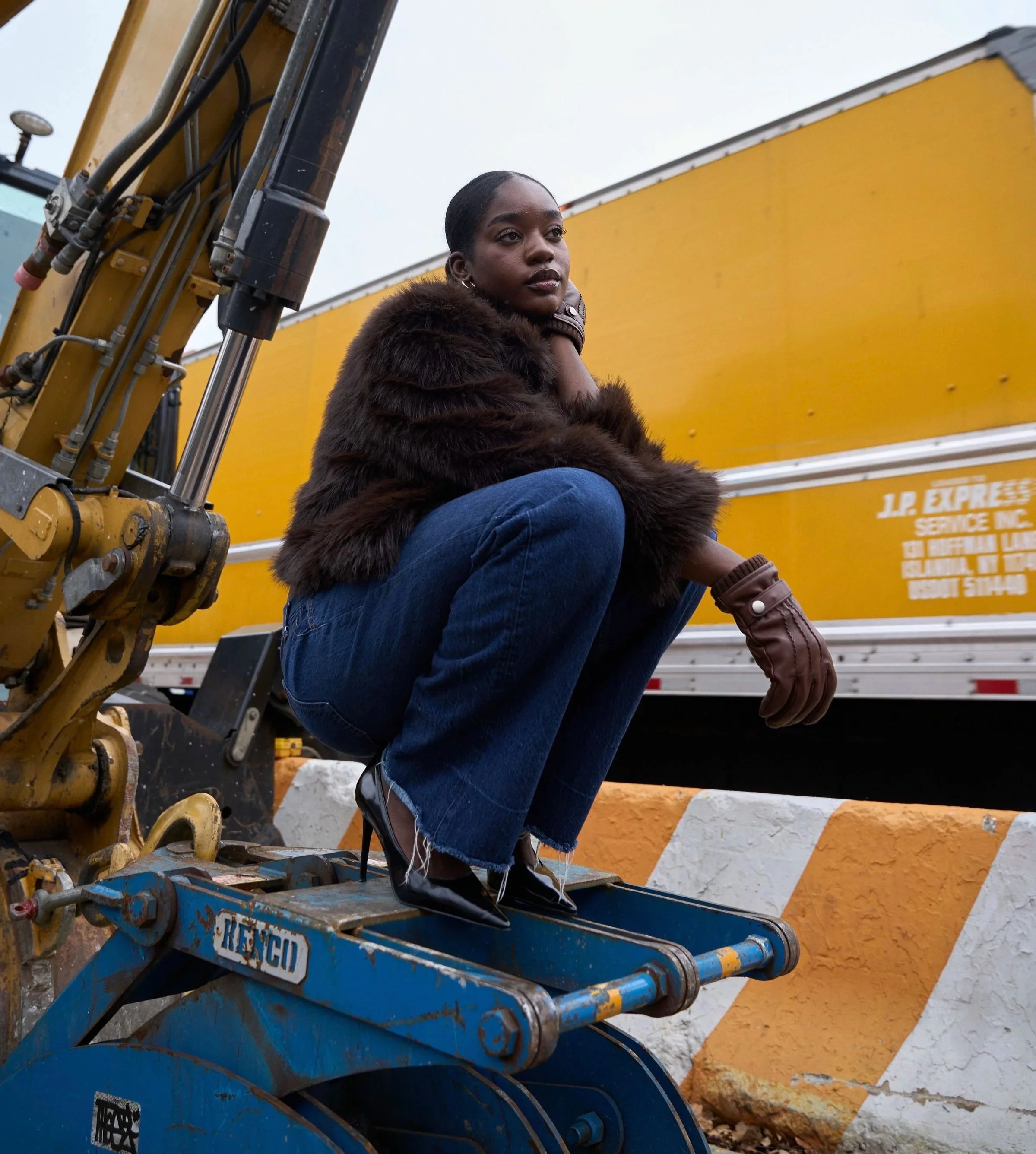 A young woman with a short hairstyle, wearing a brown fur coat, jeans, and brown gloves, is sitting on a yellow construction excavator, with a yellow truck in the background. She has a contemplative expression and is looking slightly to the side.