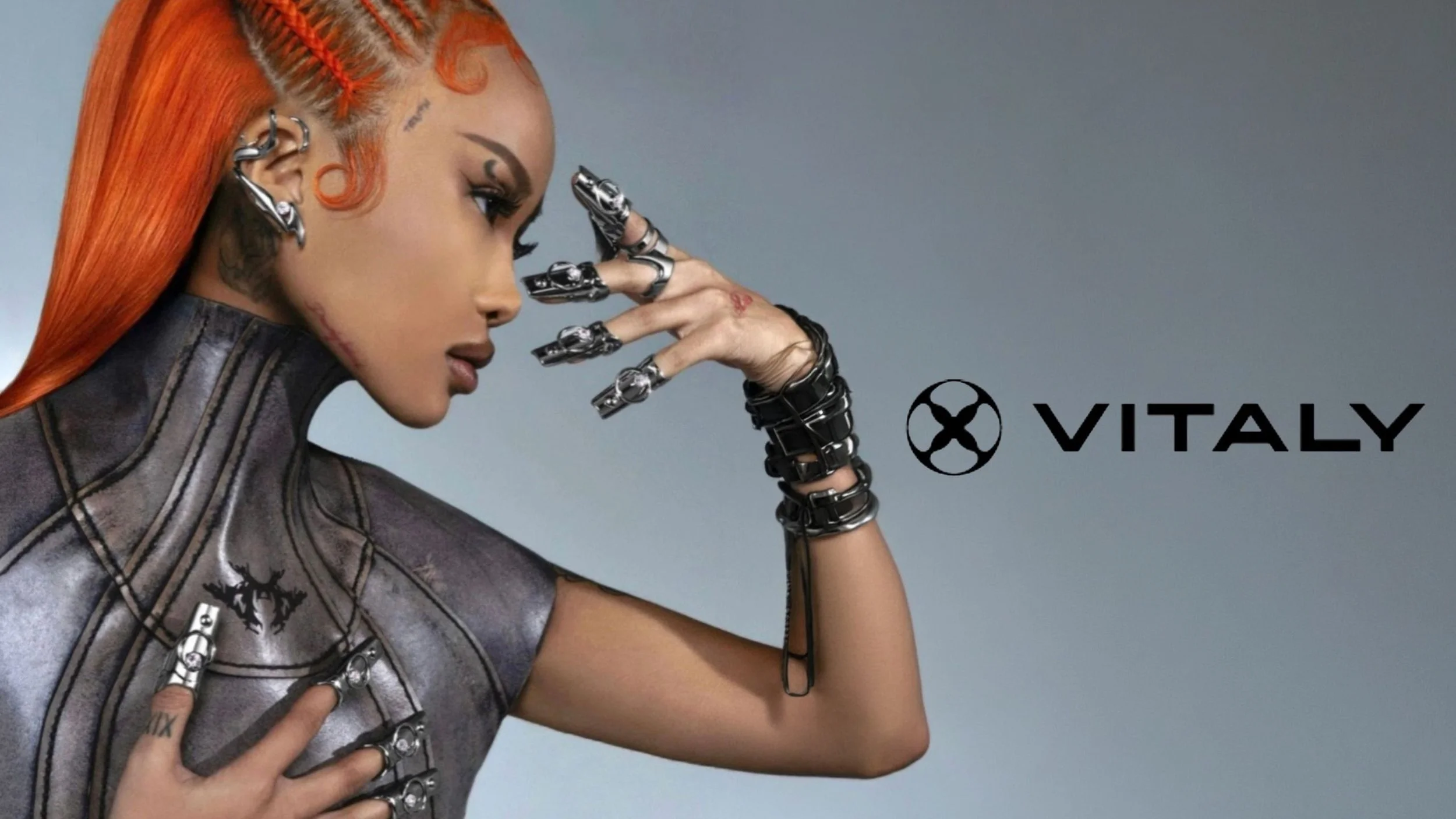 A woman with orange hair styled in braids, with tattoos on her face and hand, wearing metallic accessories and a shiny, futuristic outfit. The logo and name 'VITALY' are displayed on the right side of the image.