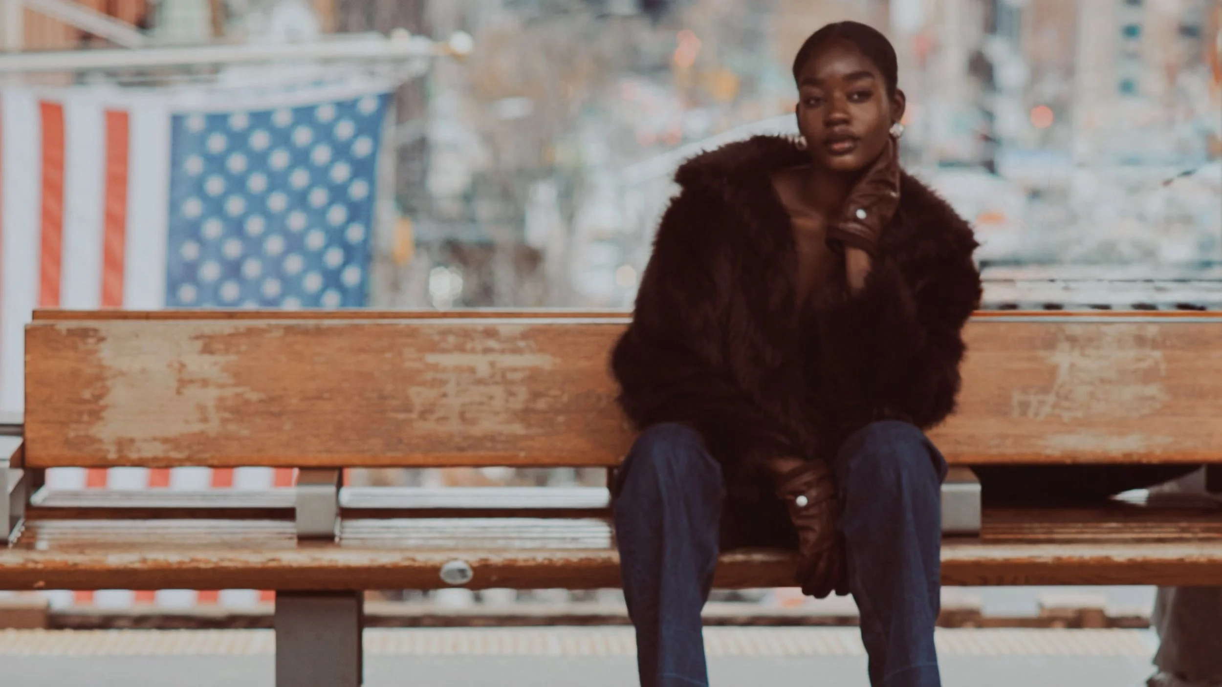 A woman with dark skin and short hair sitting on a wooden park bench, wearing a brown fuzzy coat and blue jeans, with a city street and blurred background visible behind her.