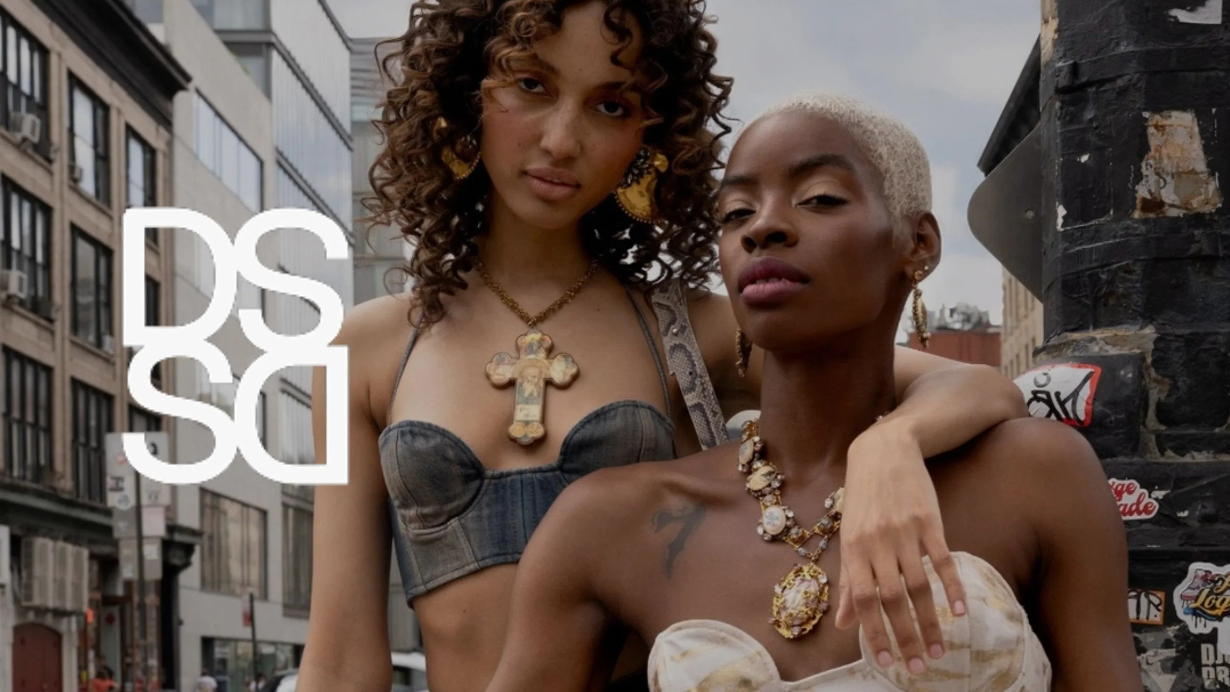 Two women with curly and short blond hair wearing jewelry and stylish clothing, standing close together outdoors in an urban setting with buildings and a cloudy sky in the background.