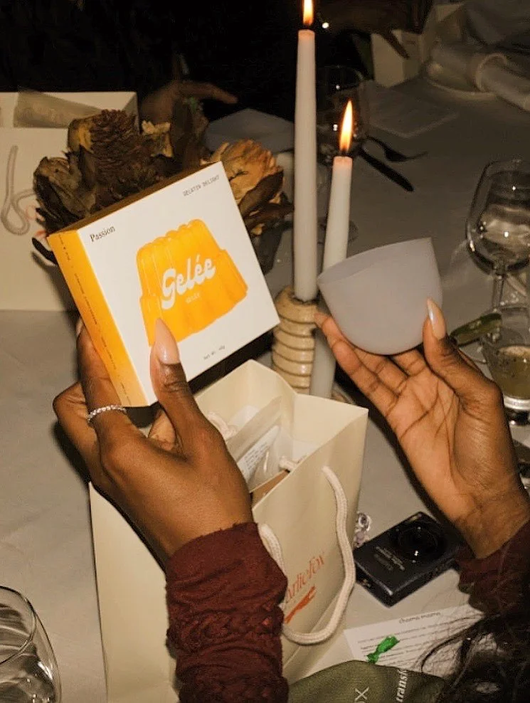 Person holding a box labeled 'Gelée' and a gray candle holder with two lit candles at a dinner table.