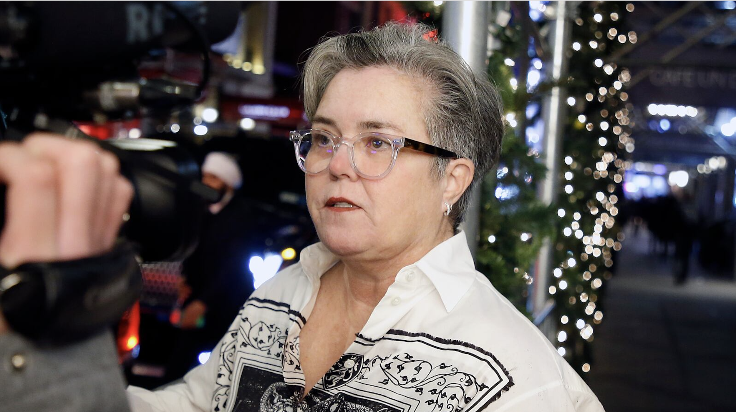 Woman with short gray hair and glasses being filmed or photographed indoors, with blurred Christmas lights and decorations in the background.