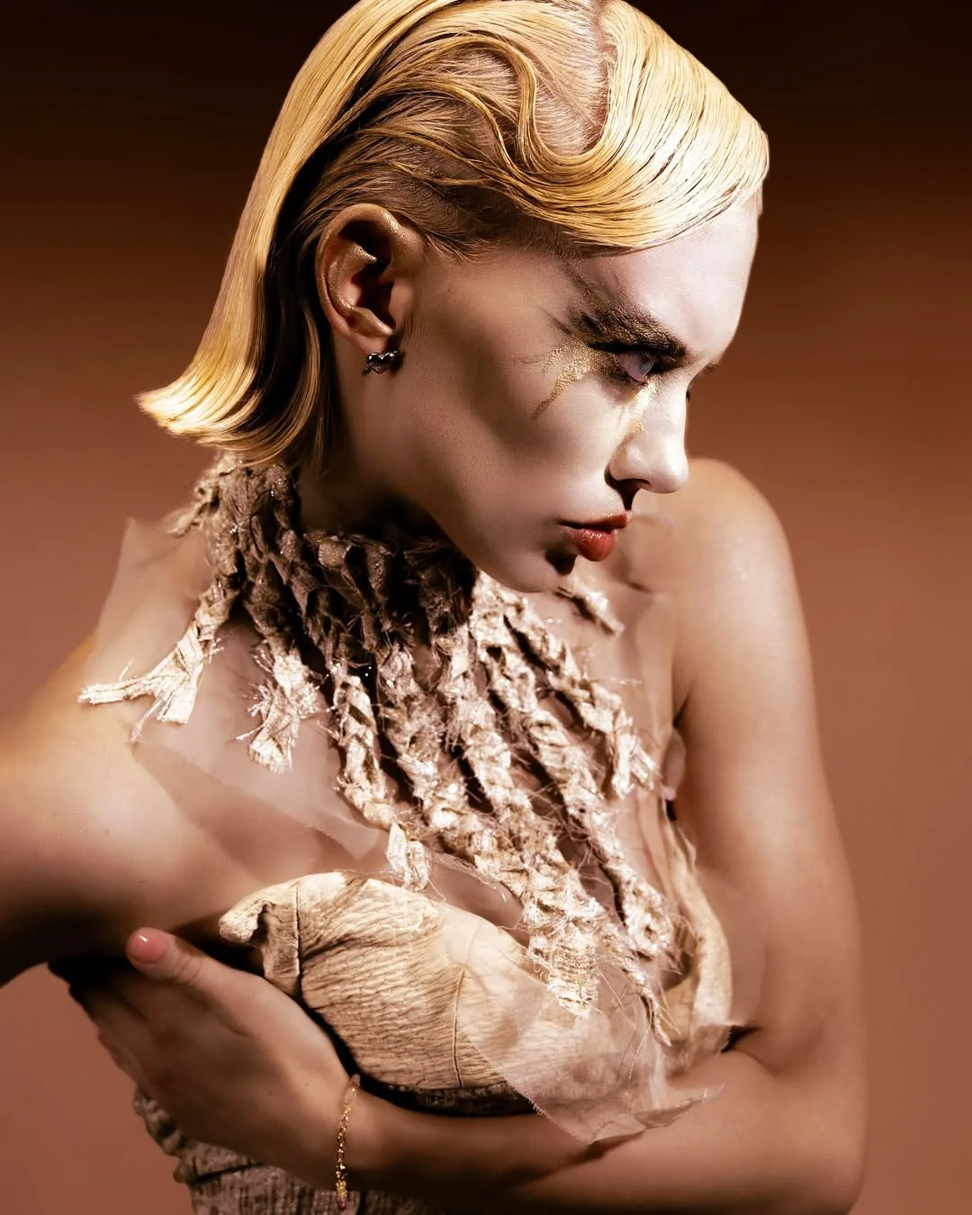 A woman with blonde, wavy hair, wearing dark earrings and makeup featuring gold accents, holds a handmade, textured clothing or fabric item against her chest. She is posed in profile with a neutral brown background.