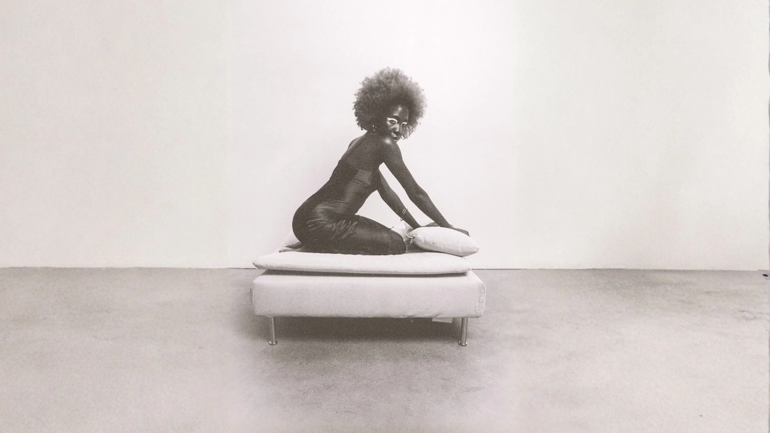 A woman with natural curly hair kneels on a small couch while adjusting a pillow in a minimalistic room with plain white walls.