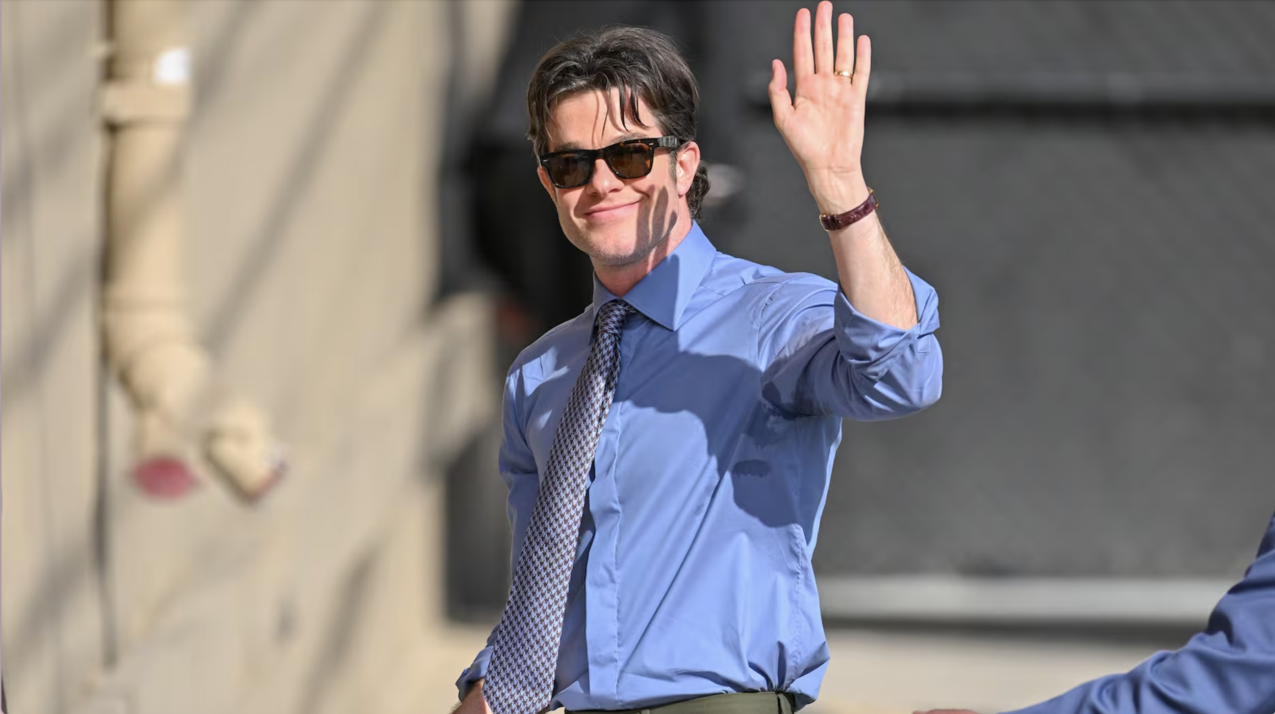 A man with dark hair wearing sunglasses, a blue dress shirt, and a patterned tie, smiling and waving
