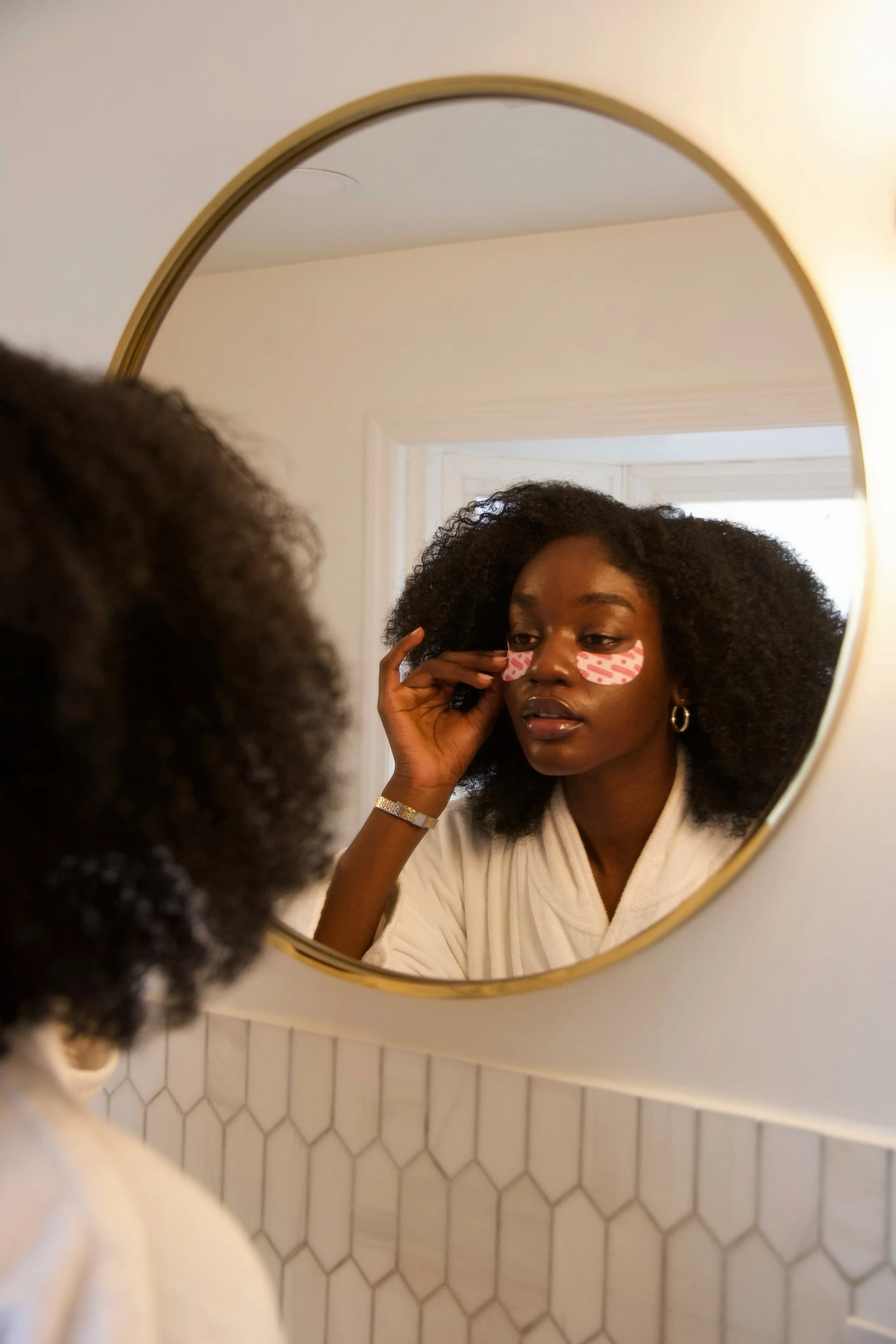 A woman with curly black hair wearing a white robe and pink face patches, looking at herself in a round mirror on a wall with white hexagonal tiles, touching her face with her left hand.