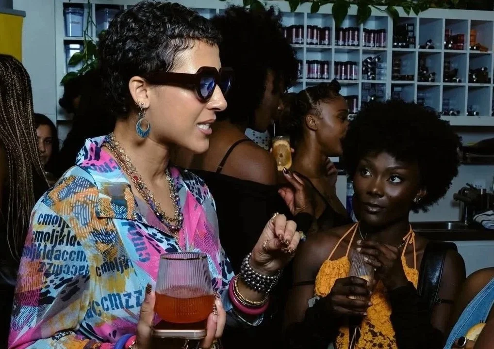 Group of women socializing indoors, one woman wearing sunglasses and colorful patterned blouse, holding a drink, others with dark skin, one with curly hair, holding a drink, in front of shelves with bottles and glasses.
