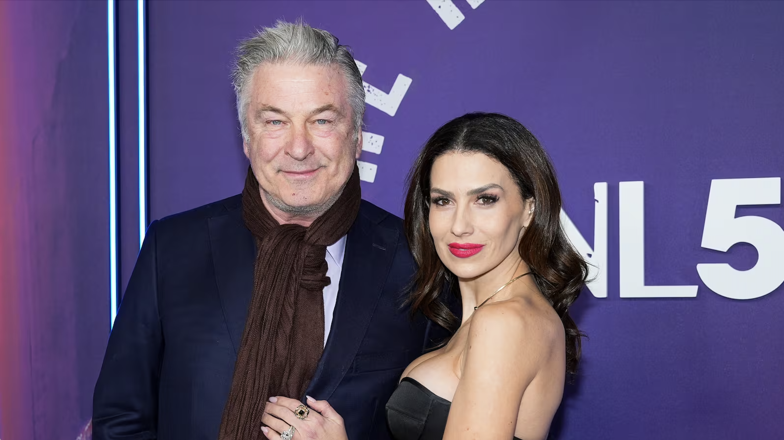 A man with gray hair wearing a dark suit and a woman with dark wavy hair in a black dress, standing in front of a purple background with white text and blue lights.