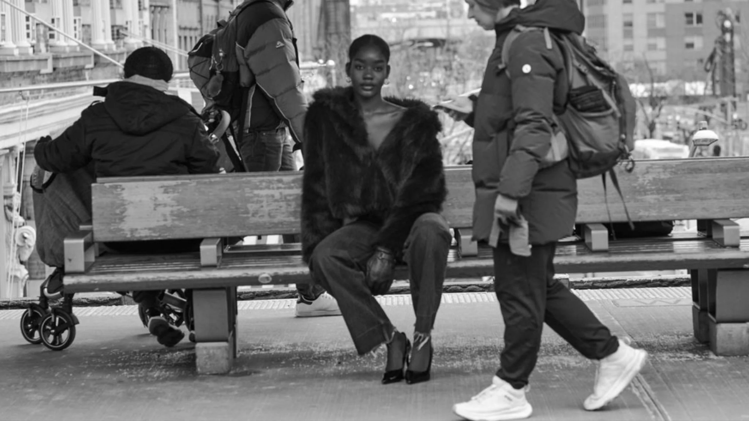 Four people in a city park. One person is sitting on a bench, another is standing and writing, the third is sitting on the bench with their head turned away, and the fourth is walking past. There is a stroller next to the bench. Modern background bui