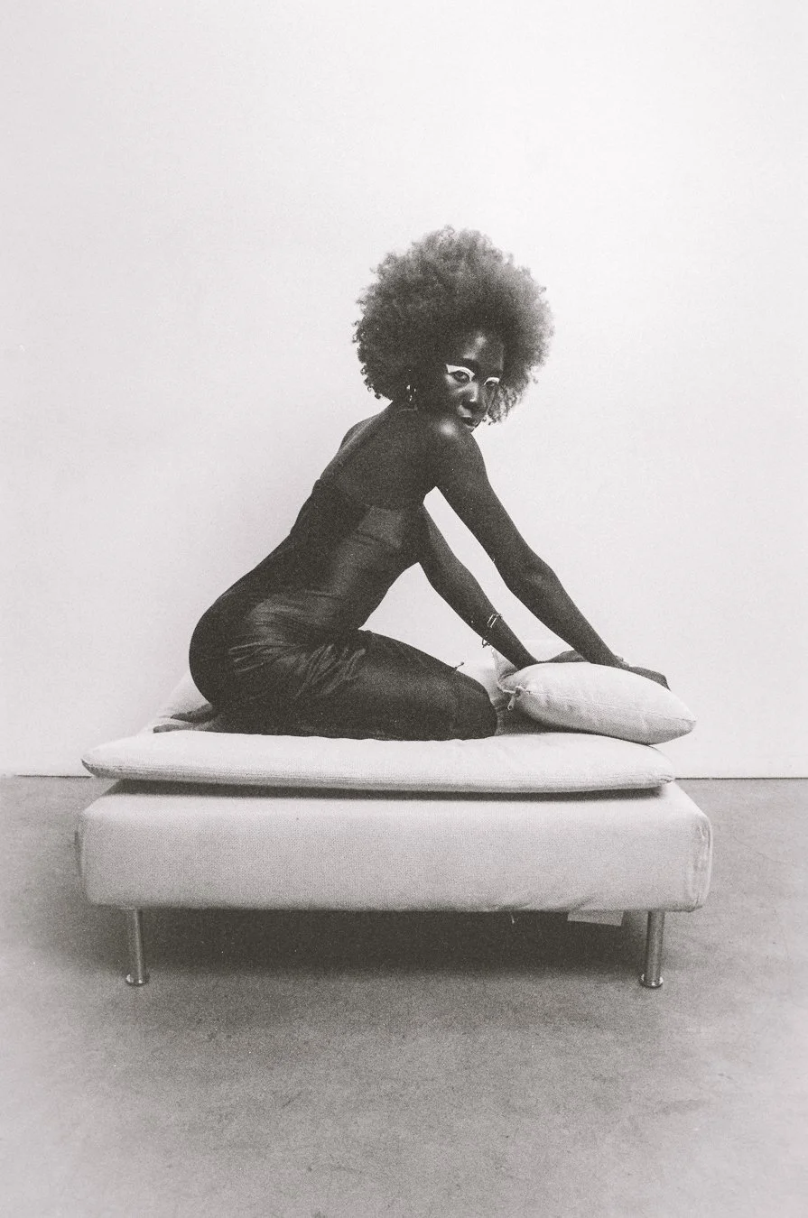 A black woman with curly hair and glasses kneeling on a bed, holding a pillow in a minimalistic room.