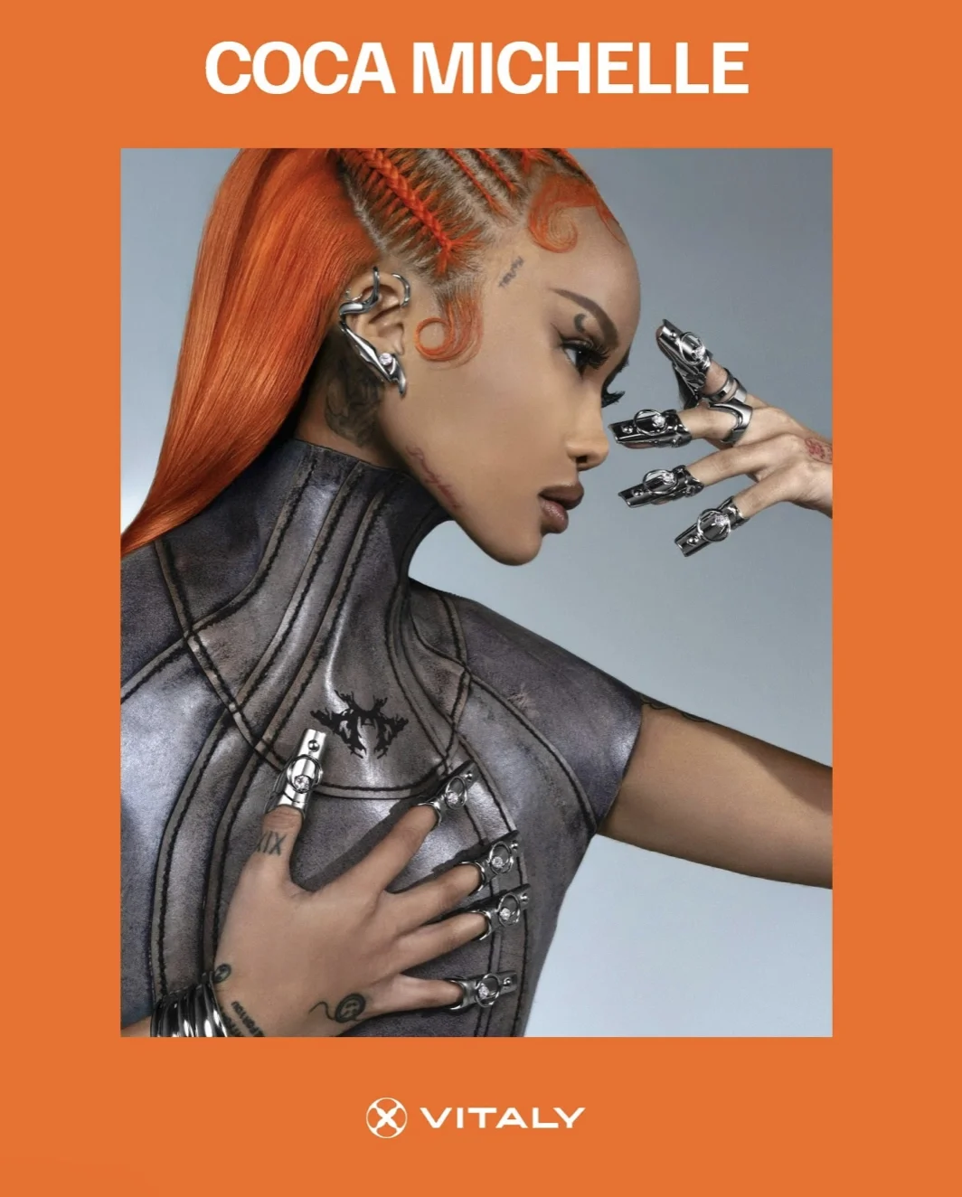 A woman with bright orange hair styled in cornrows, wearing metallic and leather clothing, and multiple futuristic rings and jewelry. The image is part of a promotional or advertising material with an orange border and the text 'COCA MICHELLE' at the top and 'VITALY' at the bottom.