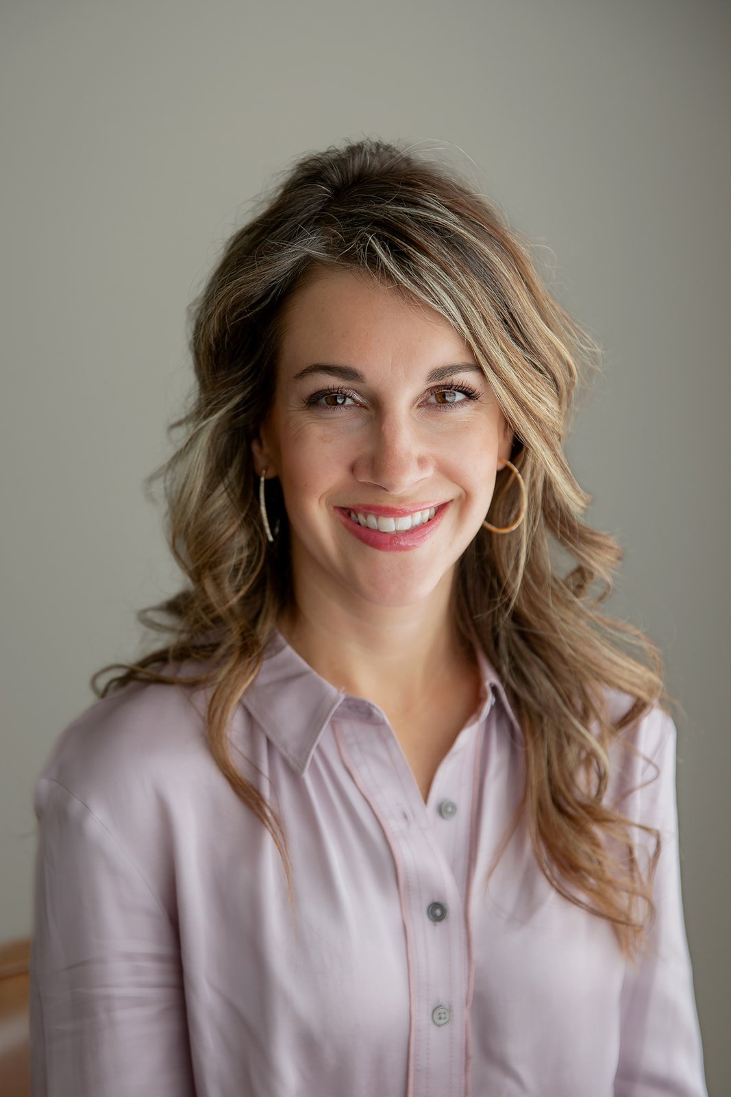 Smiling woman with wavy light brown hair, wearing hoop earrings and a light pink button-up shirt, posing indoors against a plain background.