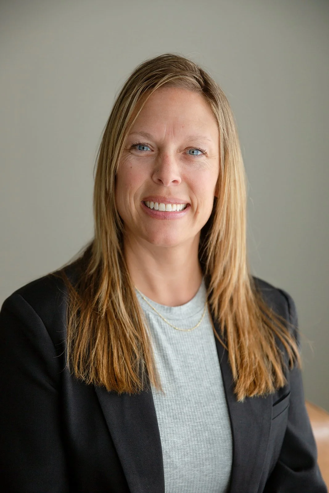 Headshot of a woman with shoulder-length blond hair, blue eyes, wearing a black blazer and light gray shirt, smiling against a plain gray background.