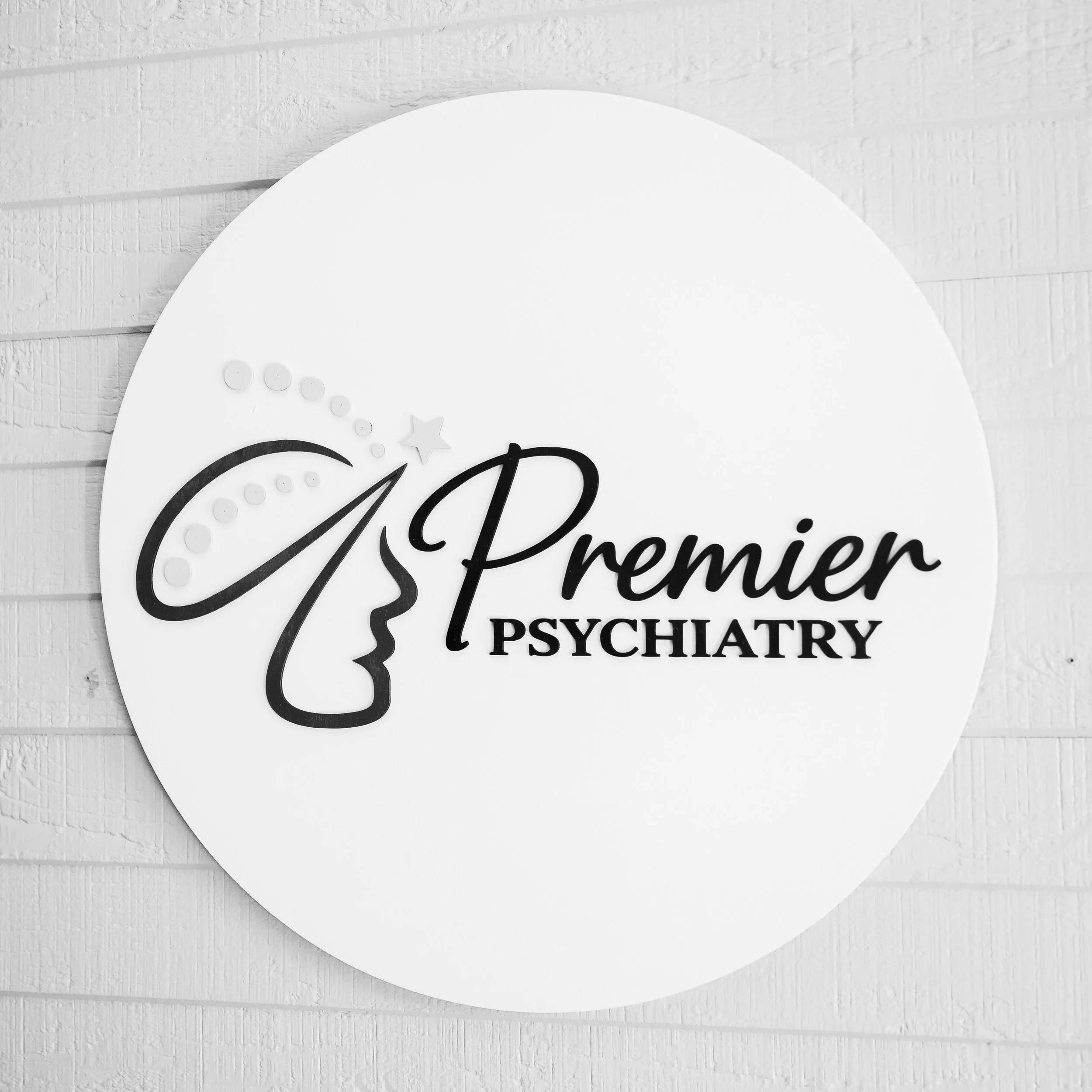 Close-up of a white wall clock with black text reading 'CSB Premier Psychiatry' and decorative stars, mounted on a white textured wooden wall.