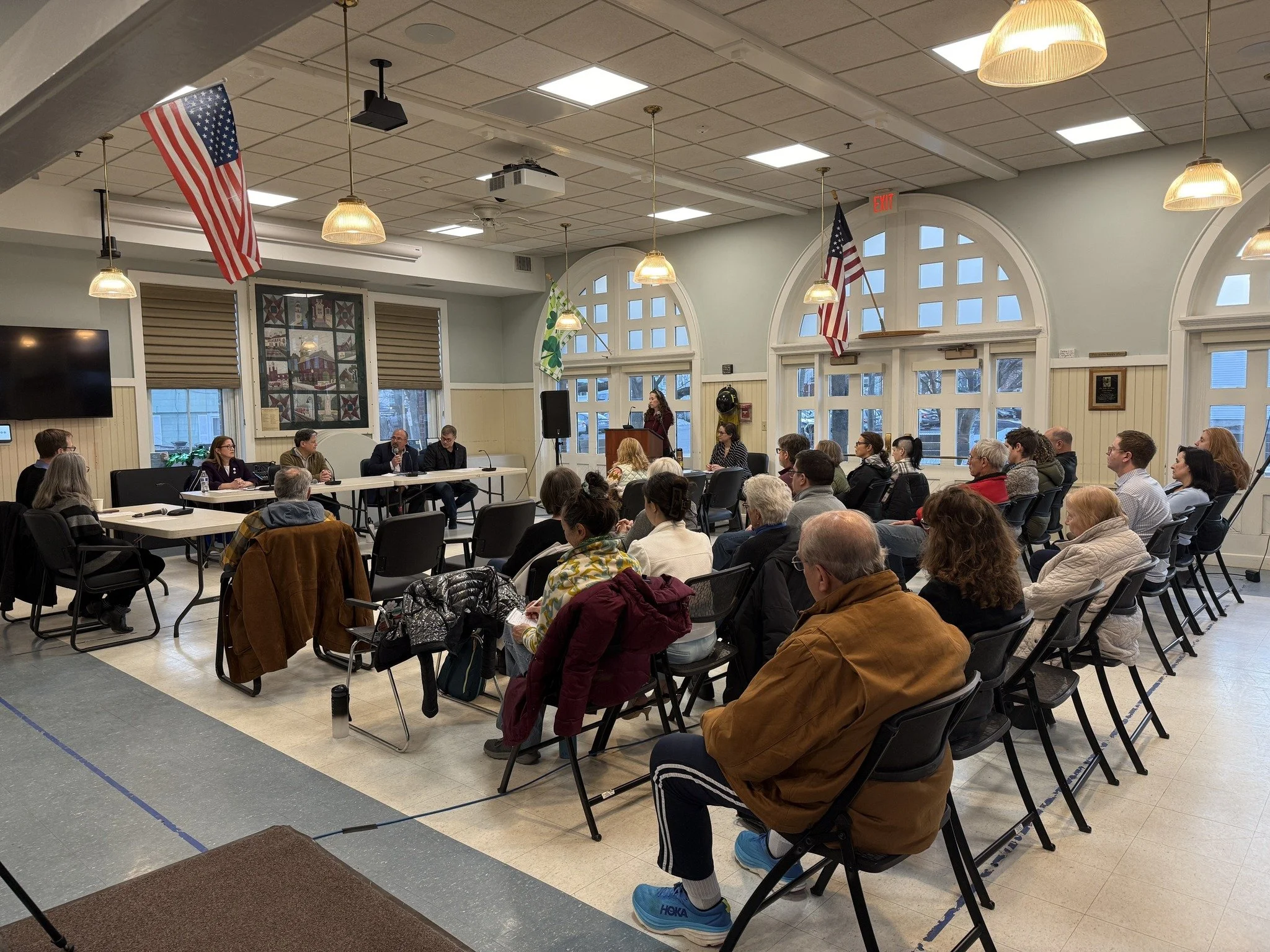 Reading Select Board Candidate Debate hosted by Reading Democrats. A room filled with people seated at tables, listening to a woman speaking at a podium and four candidates.