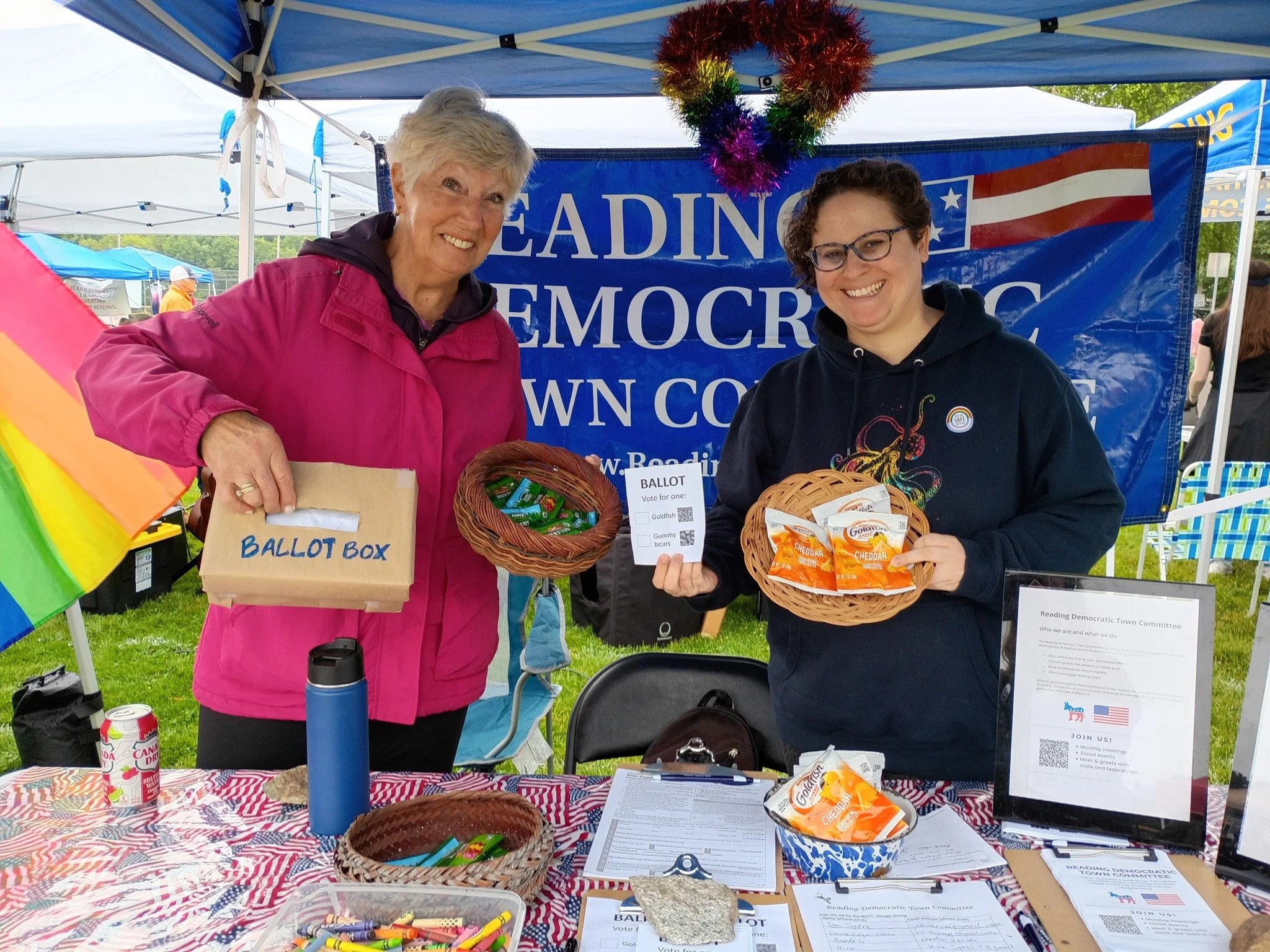 Two women at an outdoor event standing in front of a blue banner that reads 'READING DEMOCRATIC,' with a rainbow heart decoration hanging above them. The woman on the left has short gray hair and is wearing a pink jacket, and the woman on the right has curly brown hair, glasses, and is wearing a black hoodie. They are holding baskets and a ballot box, with various papers, snacks, and a rainbow flag nearby.