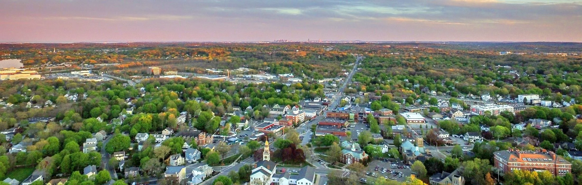 Aerial view of Reading, MA with tree-lined streets, residential houses, commercial buildings, and a church with a tall steeple, with a landscape of forests and a distant Boston skyline under partly cloudy skies.