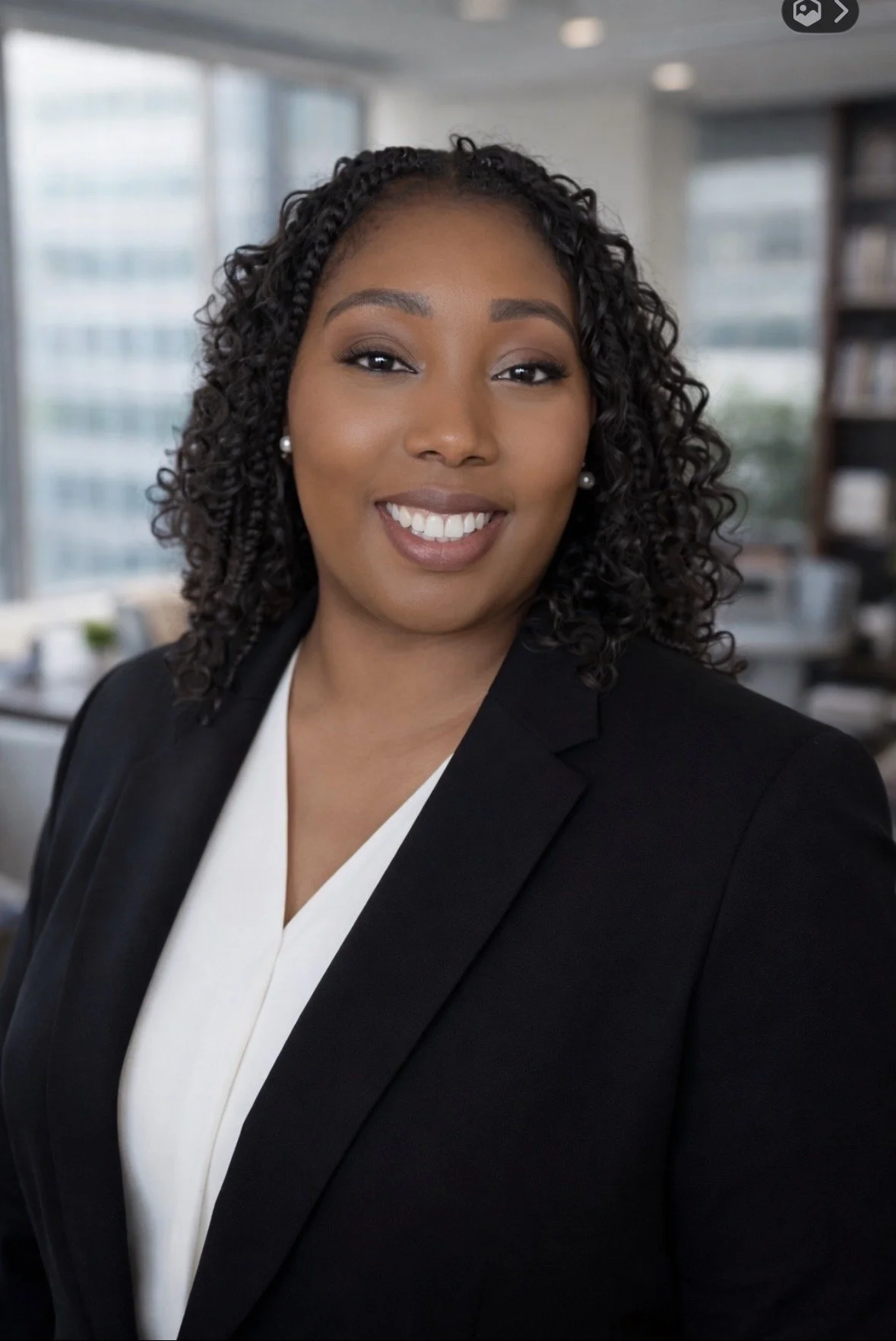 Professional woman with curly hair and earrings, wearing a black blazer and white blouse, smiling in an office setting.