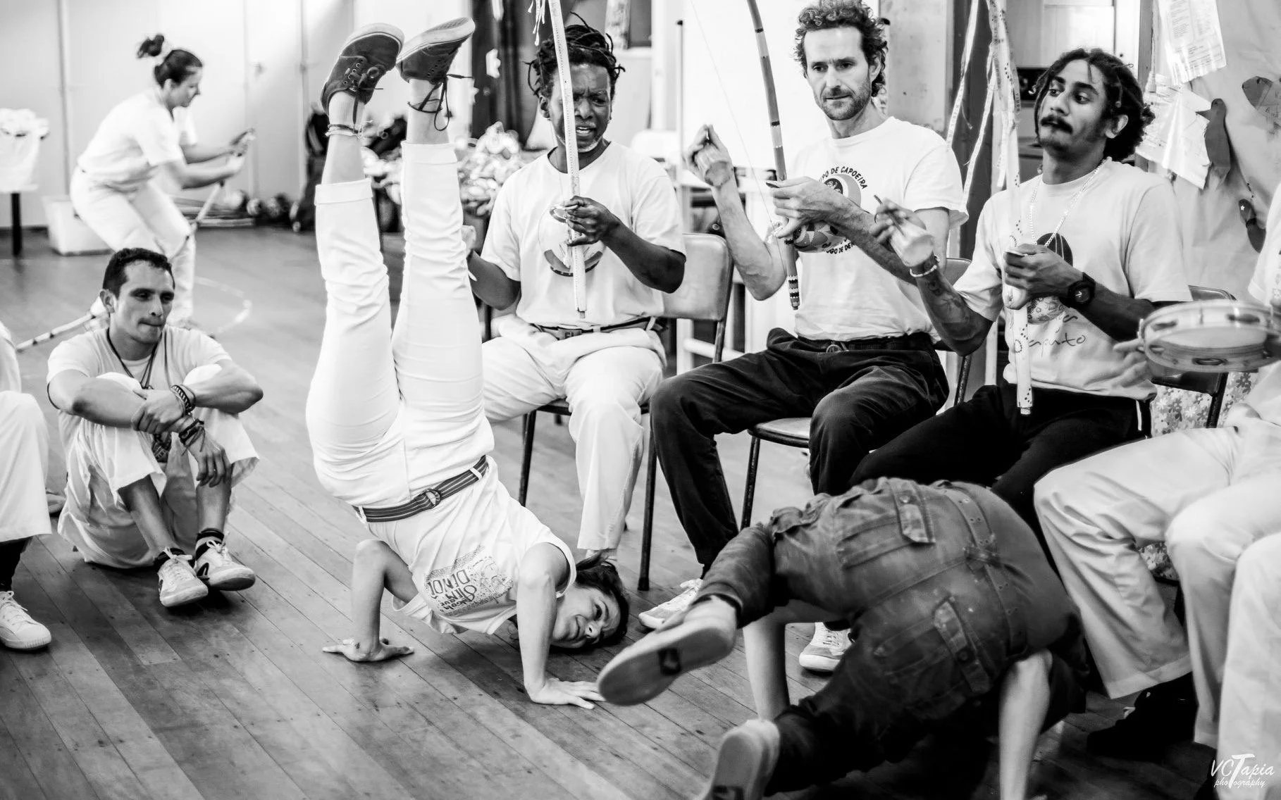 Group of people in a dance studio, some practicing acrobatic moves and others sitting or standing, with a woman doing an handstand while leaning against a person in front of her.