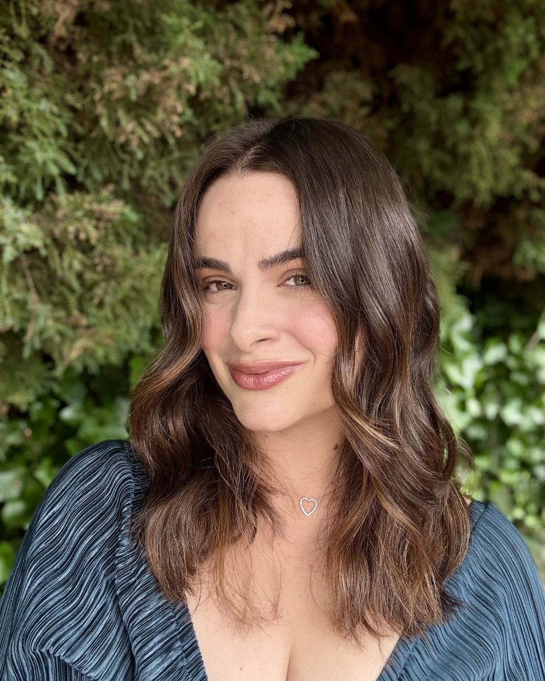 A young woman with wavy brown hair and light skin, smiling slightly, wearing a black top and a silver heart necklace, standing in front of green foliage.