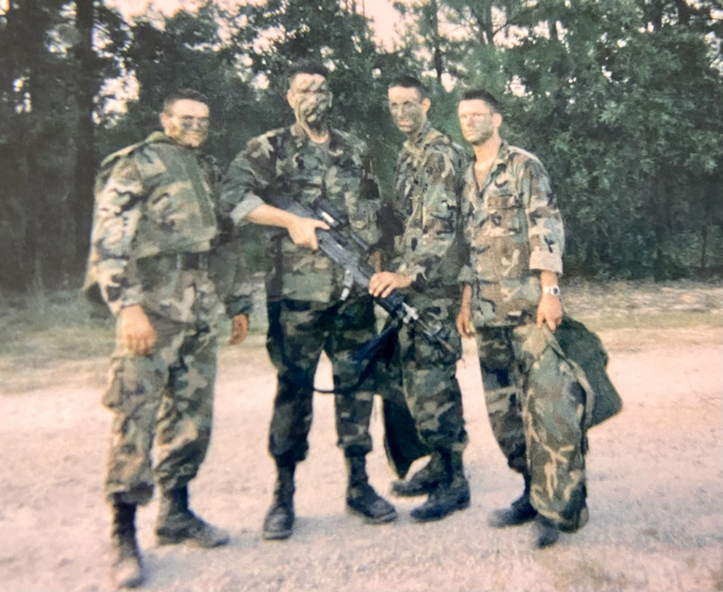 Four soldiers in camouflage uniforms standing outdoors in front of trees and bushes during dusk or dawn. Secretary of War Pete Hegseth.