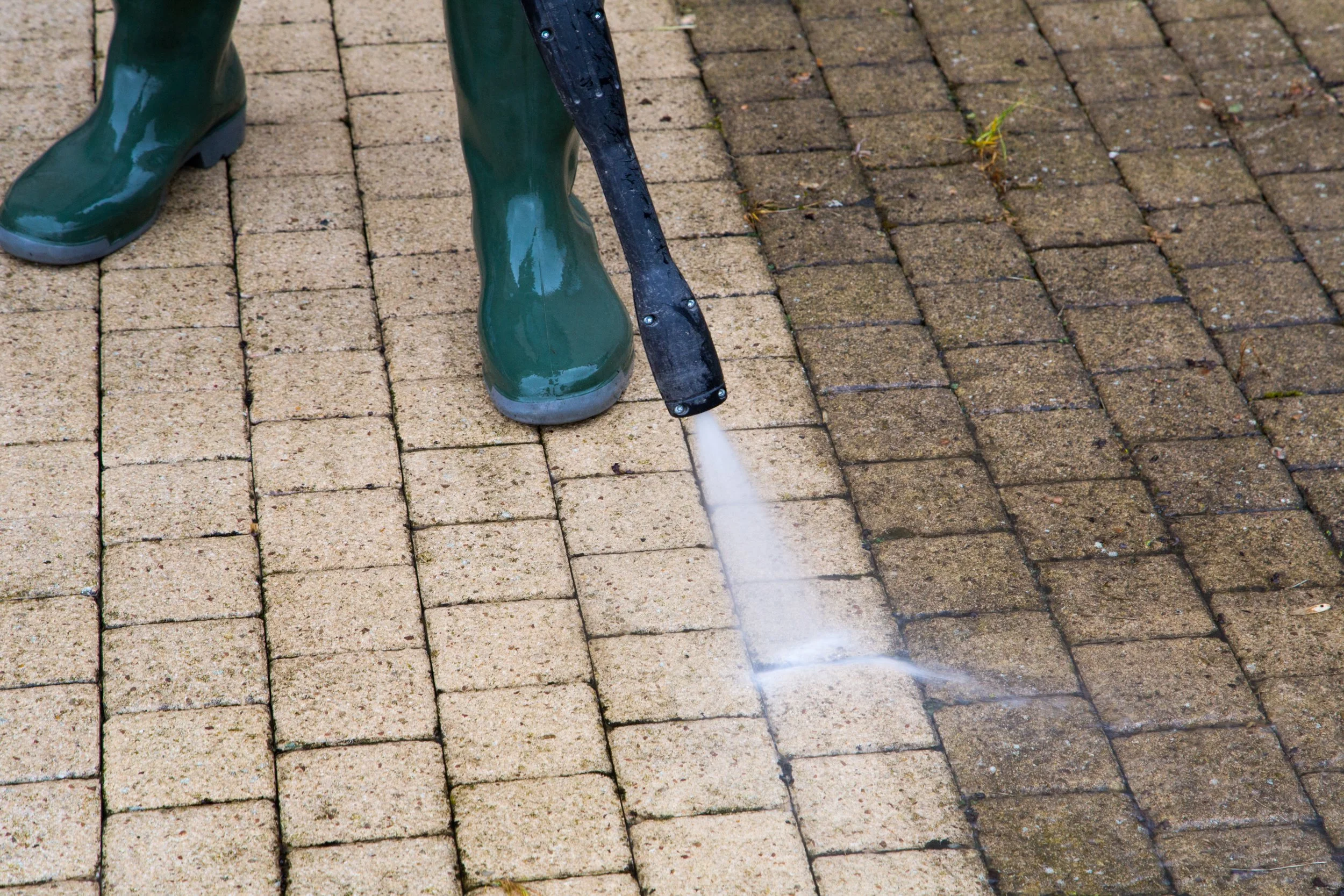 Una persona usando botas verdes y una herramienta de inmunización rociando agua sobre un pavimento de ladrillos.
