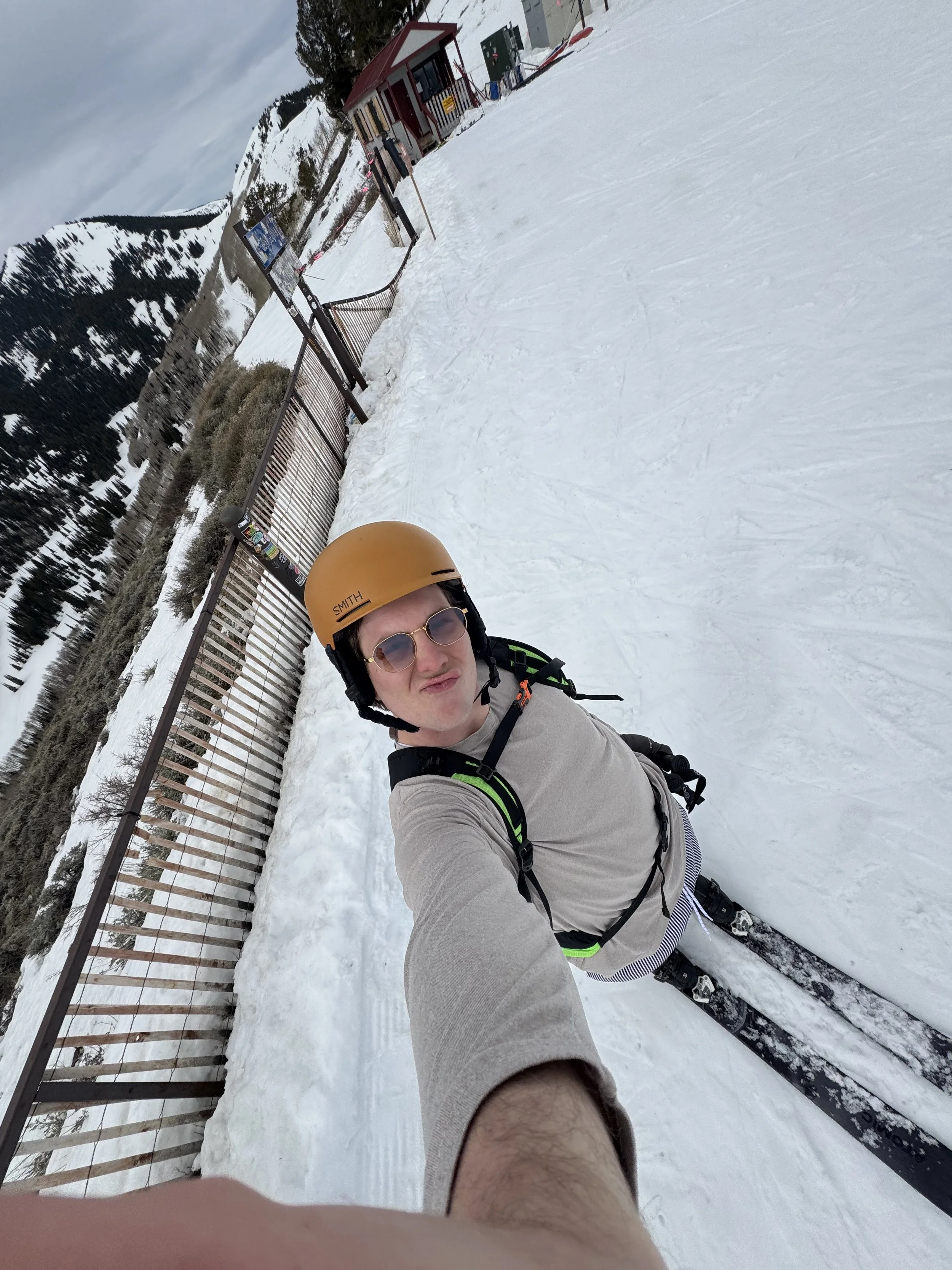 A person wearing an orange helmet, sunglasses, and a gray shirt taking a selfie on a snowy mountain with ski equipment. In the background, there is a small building, a fenced area, and snow-covered trees and mountains under cloudy skies.