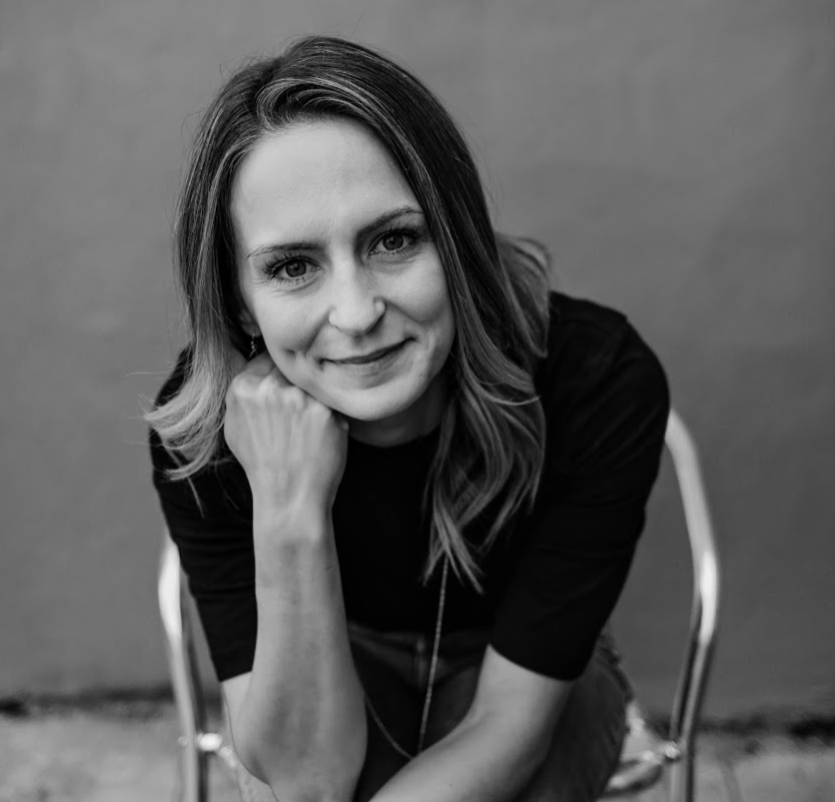 A black and white portrait of a woman with shoulder-length hair, smiling slightly, resting her chin on her hand, sitting on a chair against a plain background.